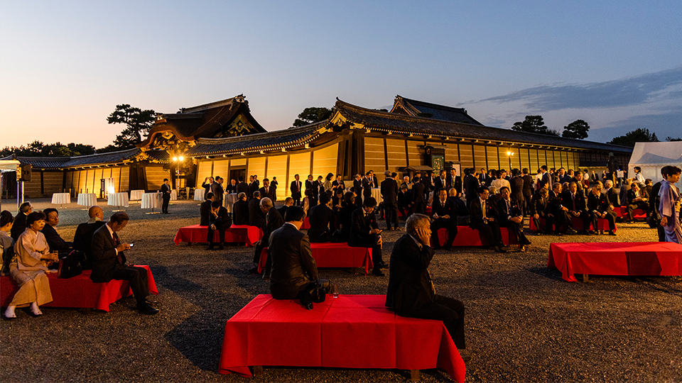 Business event in front of Japanese castle and Unesc world heritage site, Nijo-jo castle with wooden walls and intricate entrance gates. Attendees sit on red-cloth draped benches.