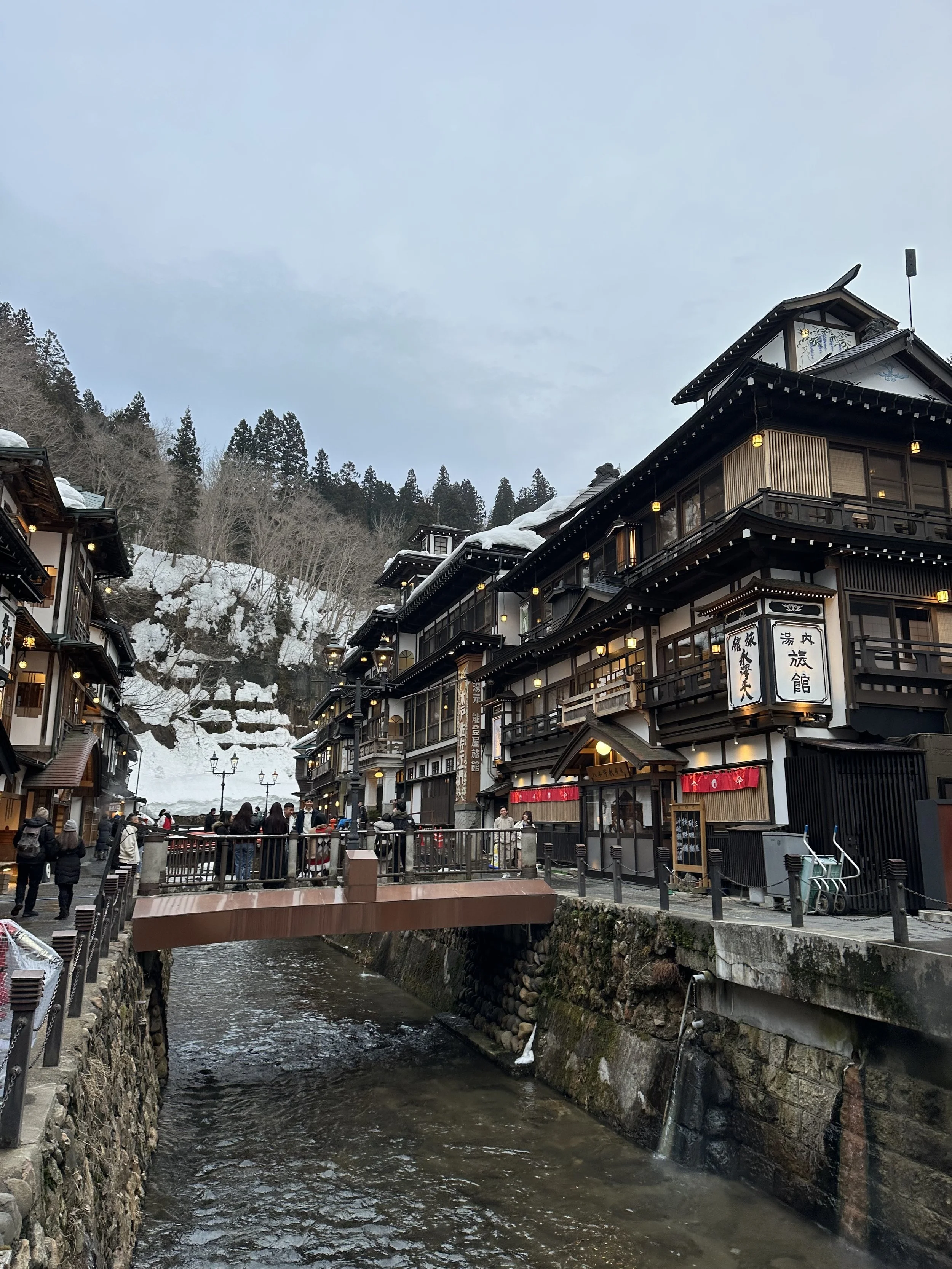 A bustling street scene in a snowy mountain area with traditional Japanese buildings lining a small river, and people walking around.