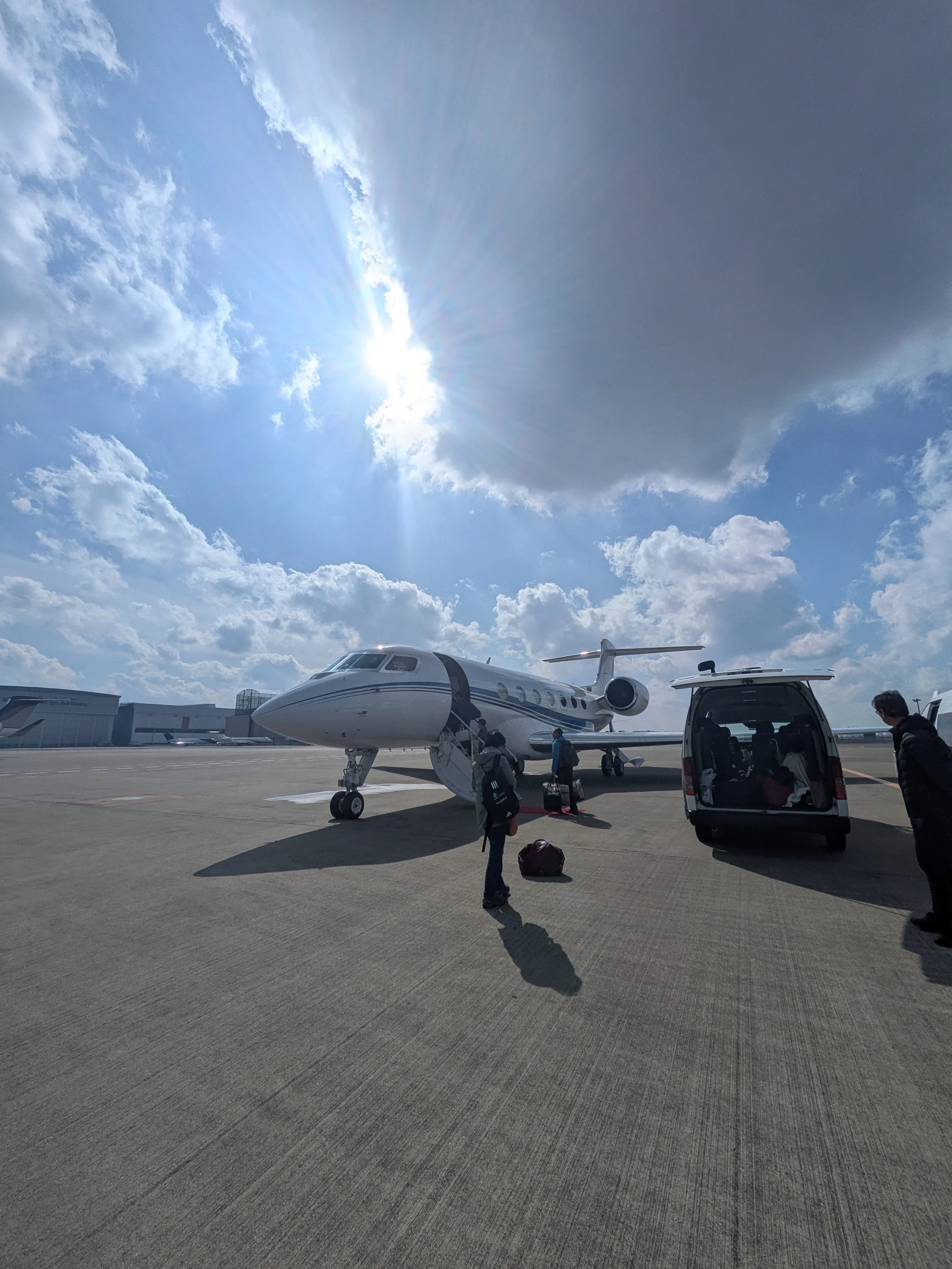 People boarding a private jet on an airport tarmac under a partly cloudy sky with the sun shining.