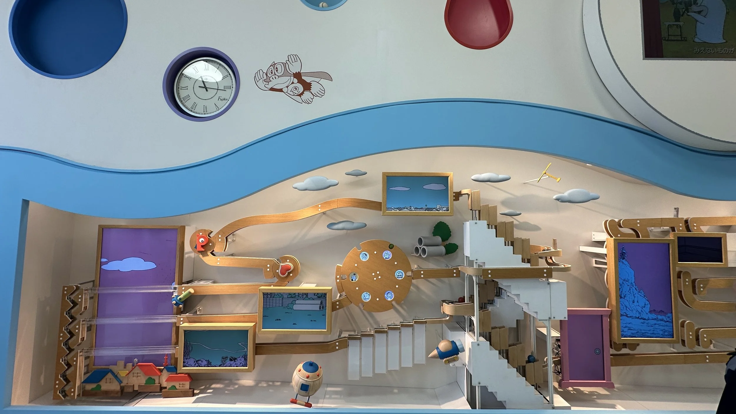 A whimsical wall with a clock, curved colorful shapes, wooden blocks and structures that all interact with eachother as a ball rolls through the a track and triggers chain reactions among the sculpture.