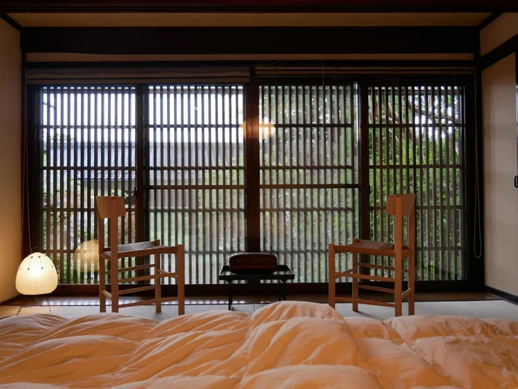 Image of a wooden sliding door with slated panels, two wooden chairs in front of it and an egg shape lamp next to one chair with a white down comforter  seen from the foreground. Warm lighting is throughout the picture.