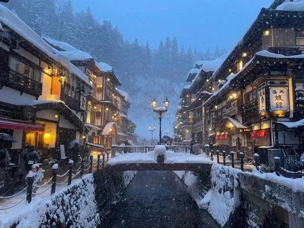 A Japanese onsen town with traditional 3-story wooden architecture along a stream with a foot bridge lit by the dusk light and street lamps while snowing in Ginzan Onsen, in Yamagata Japan.