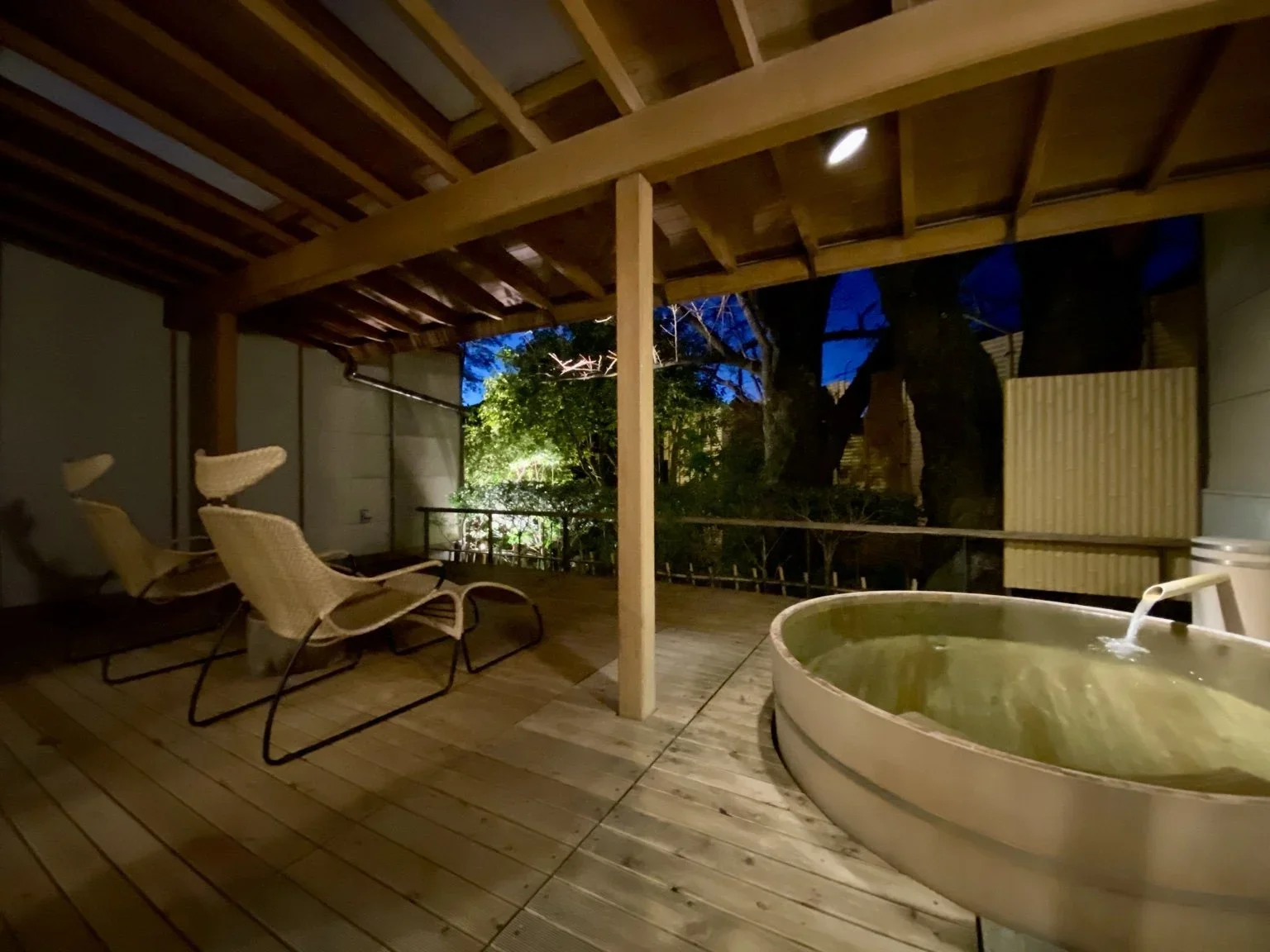 Image of a private wooden patio with two chairs, a wooden bath overlooking a small green garden with bushes and  big tree at dusk.