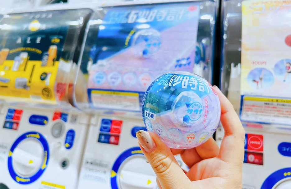 A hand holding a round ball in front of a series of stacked gacha machines, or machines that you crank the handle and win a random toy in a ball.