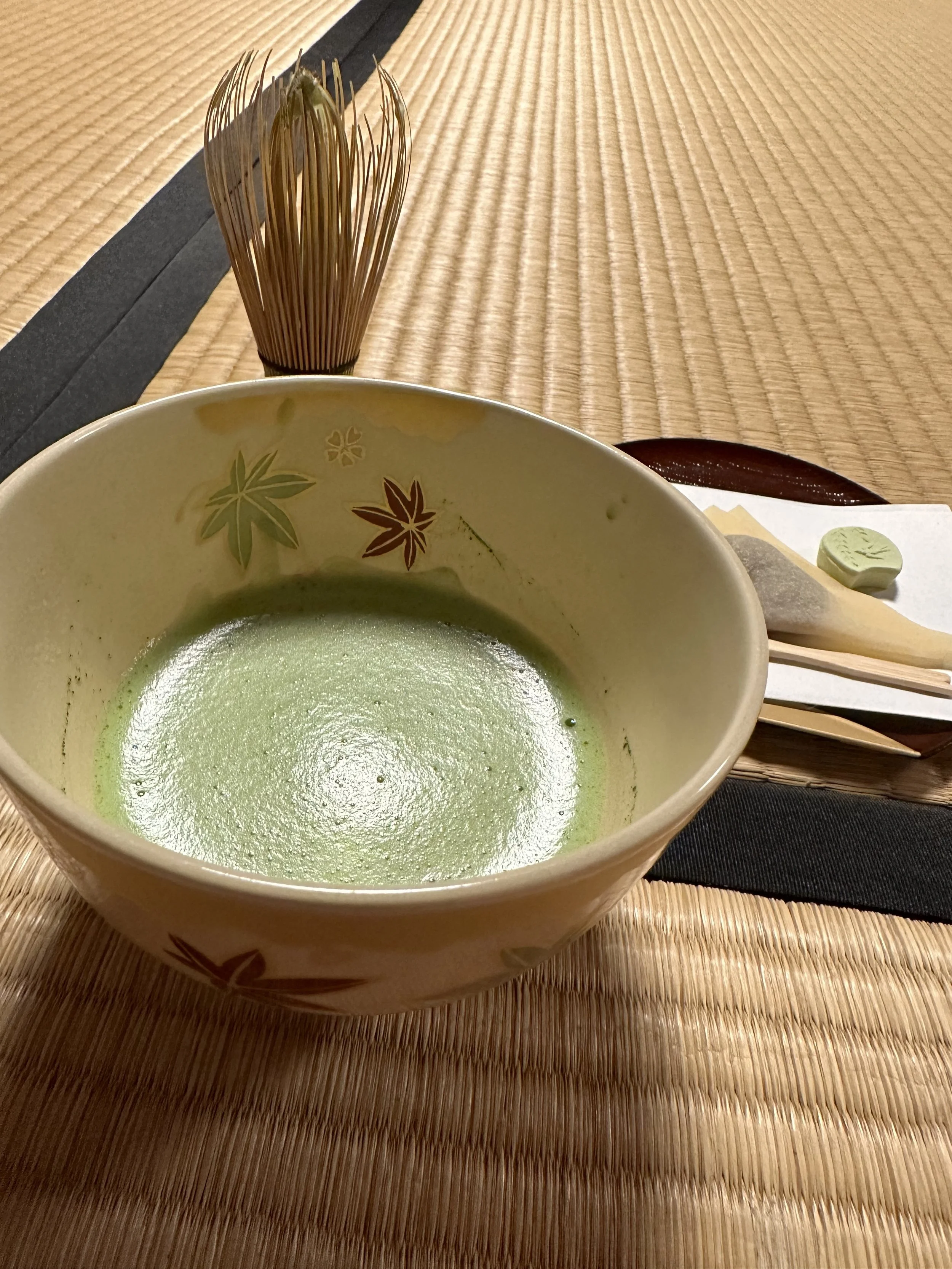 Bowl of freshly frothed matcha sits on a tatami mat floor with bamboo whisk and Japanese sweets on a plate behind the bowl