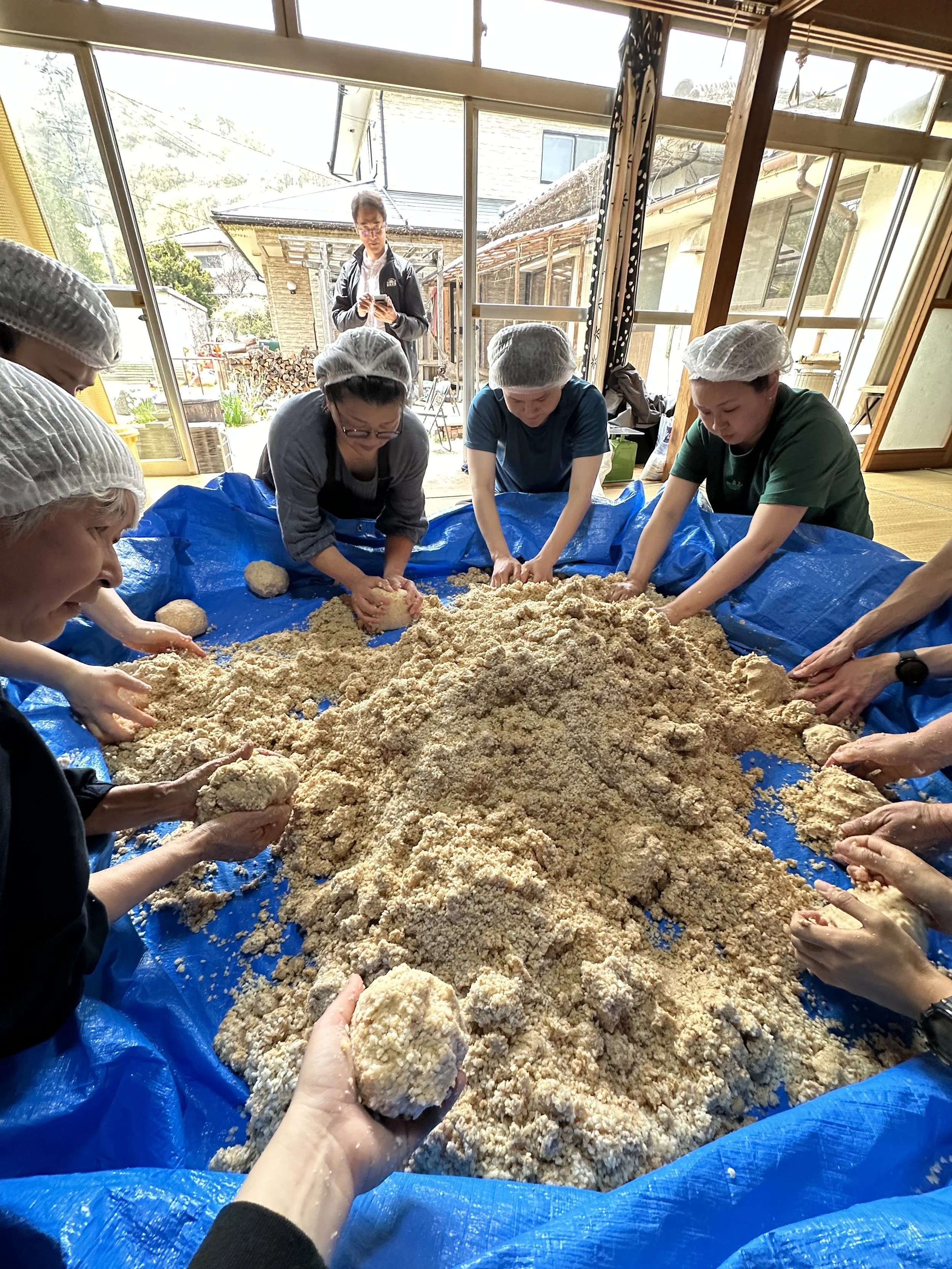 People kneading dough or a similar substance on a large blue tarp inside a bright room with large windows, with a person standing outside taking a photo.
