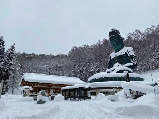 A snow covered seated Buddha in a tree lined landscape.