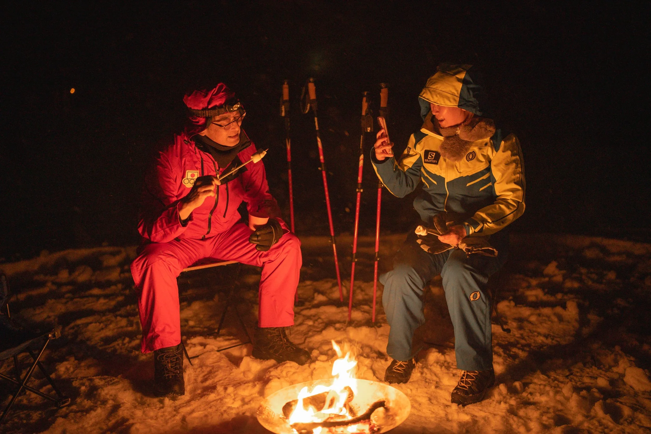 Two people dressed in winter outdoor gear sitting around a campfire on snow at night, with skis and poles standing behind them, one holding a phone and the other holding marshmallows for roasting.