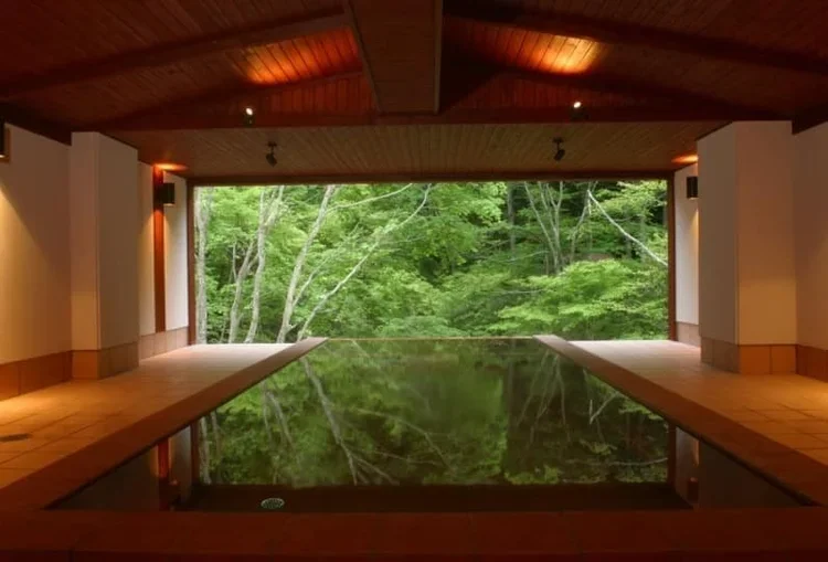 An onsen pool reflecting the lush green garden through large open windows inside a wooden room with warm lighting in matsumoto, japan.