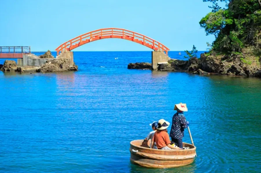 People in a small round boat floating on blue water near a rocky shoreline with a bright orange bridge overhead and lush green trees on the right side.