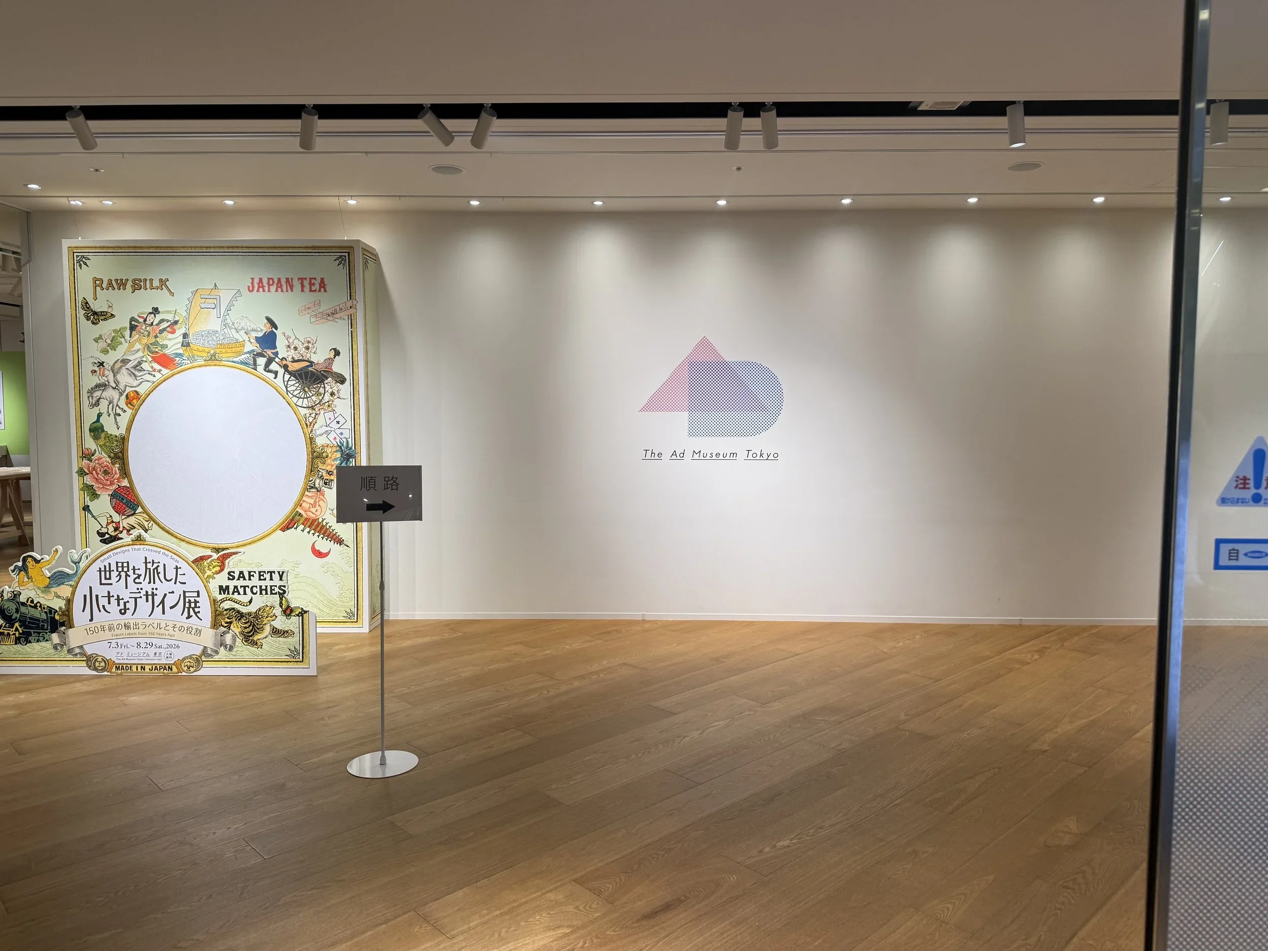 Stark entrance of the Ad museum with the logo on a white wall and a wooden floor and a small display of the exhibition.
