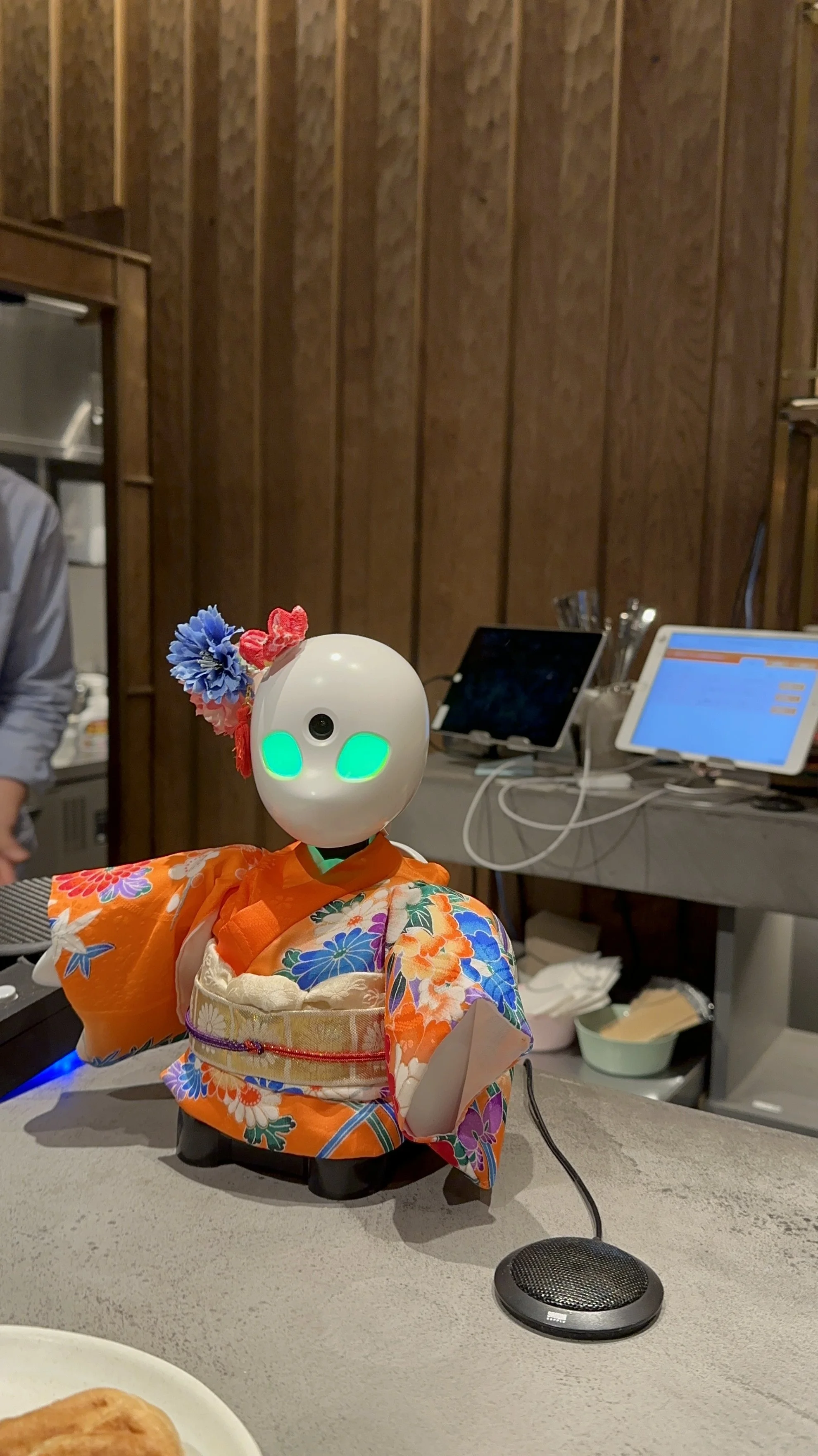 Image of a solo mini robot with glowing green eyes and a white face with flowers on the top of its head on the counter at a cafe.
