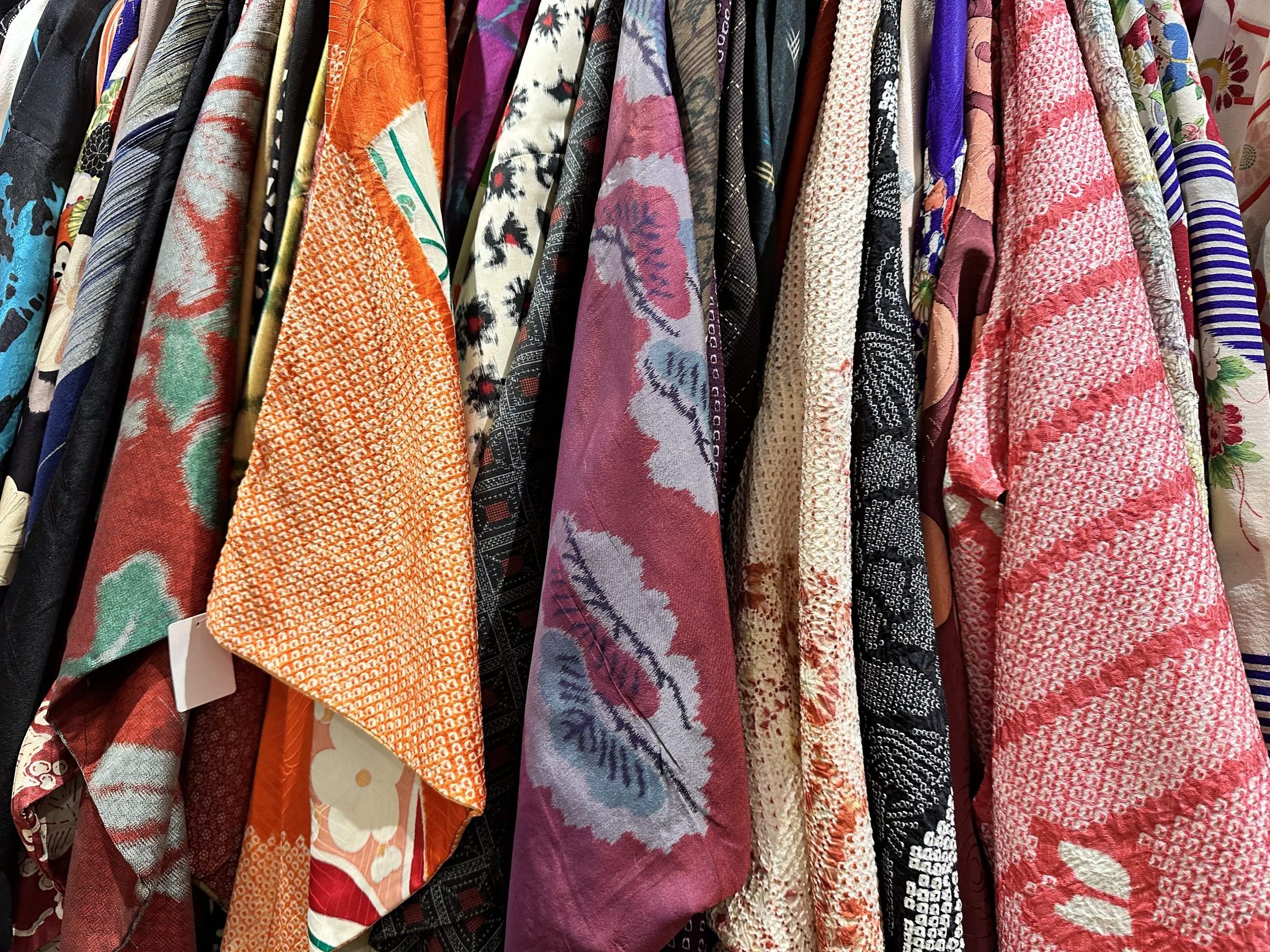 Different styles of kimonos hand side by side including meisen and shibori hang in a second-hand shop.