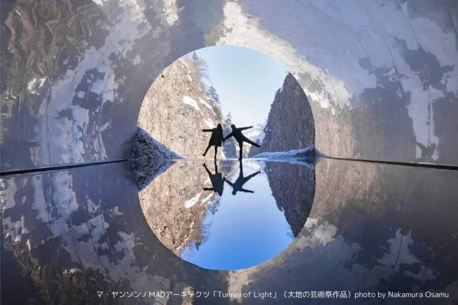 Two people stand in tunnel with a winter landscape at the end reflecting in a pool of water.