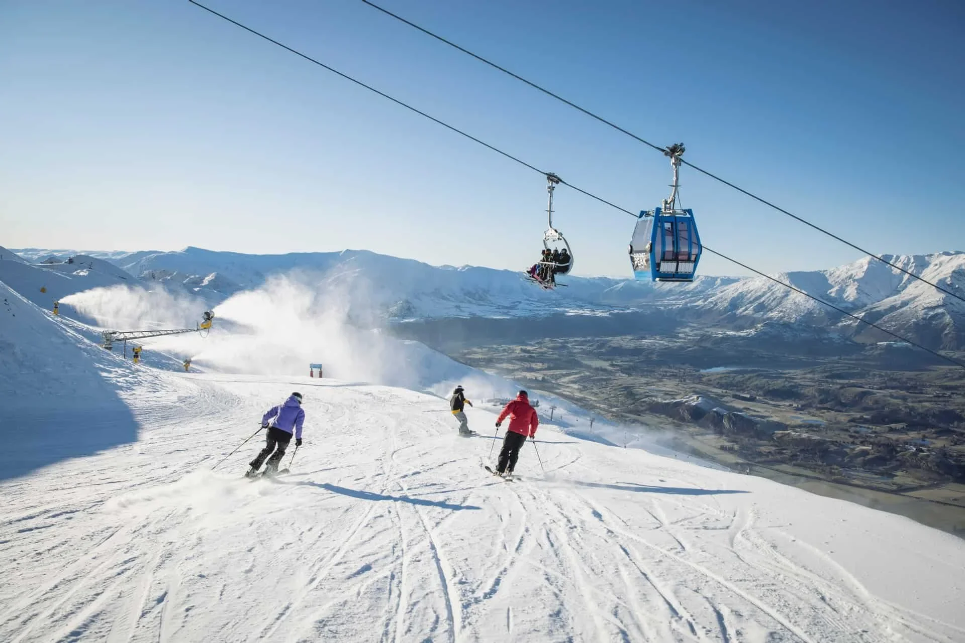 Skiers on a snowy mountain slope with ski lifts in the background, mountains in the distance, and a clear blue sky.