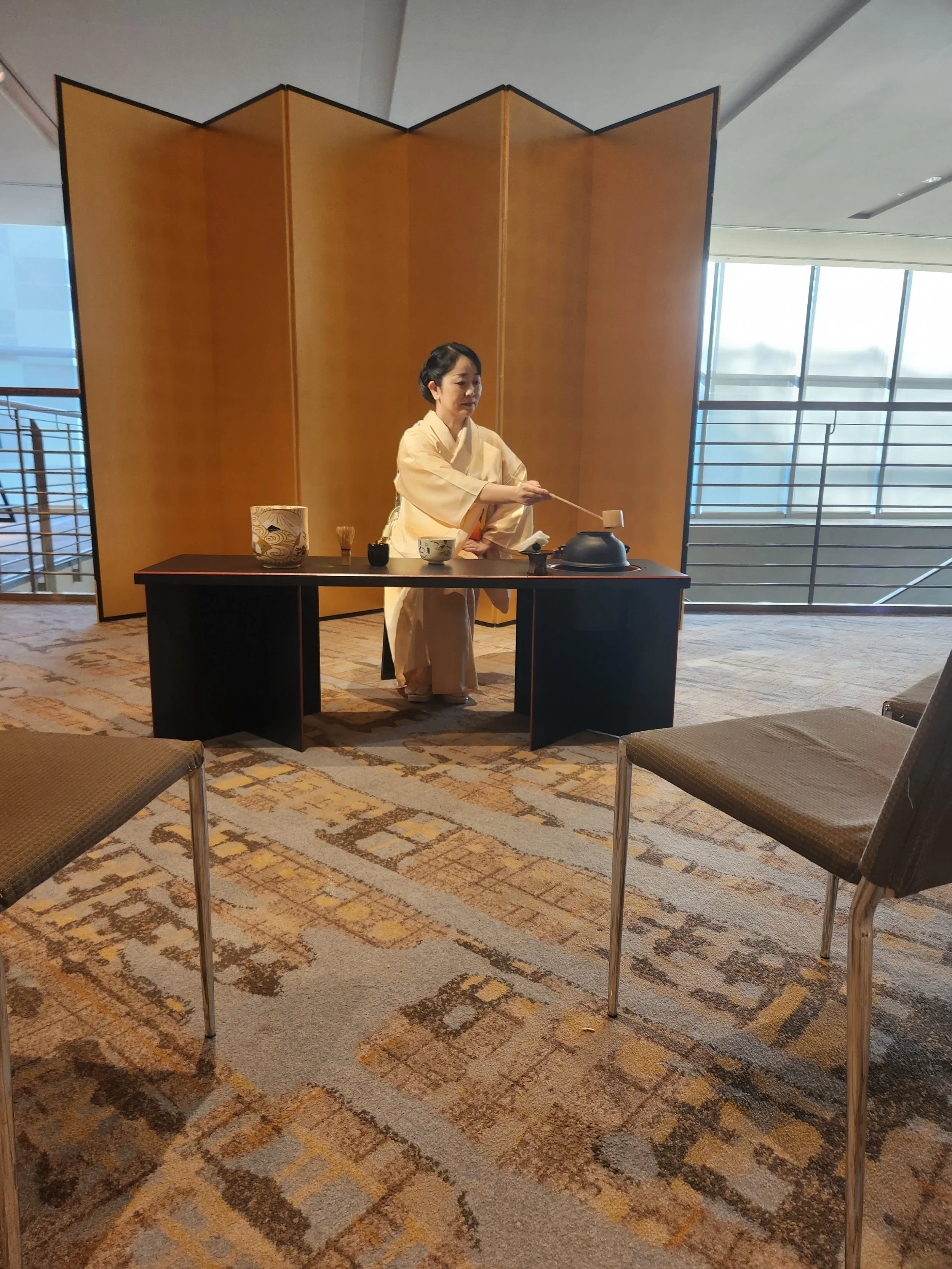Woman in a kimono is holding a tea ceremony ladle at a low table in front of a golden folding screen in a hotel ballroom in Kyoto at a corporate event.