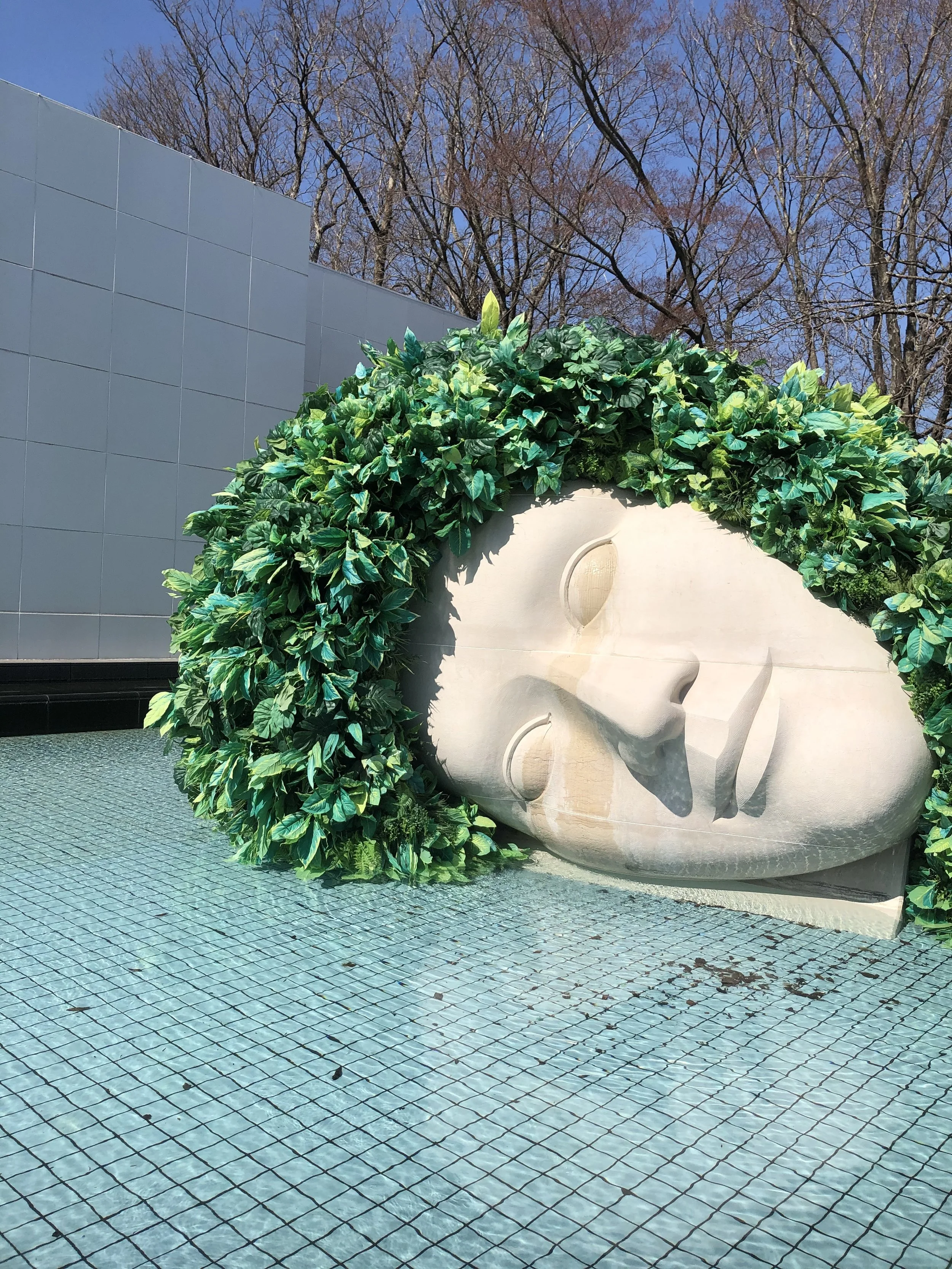A large white sculpture of a sleeping face surrounded by greenery, with a blue tile fountain in the foreground and leafless trees and a blue sky in the background.