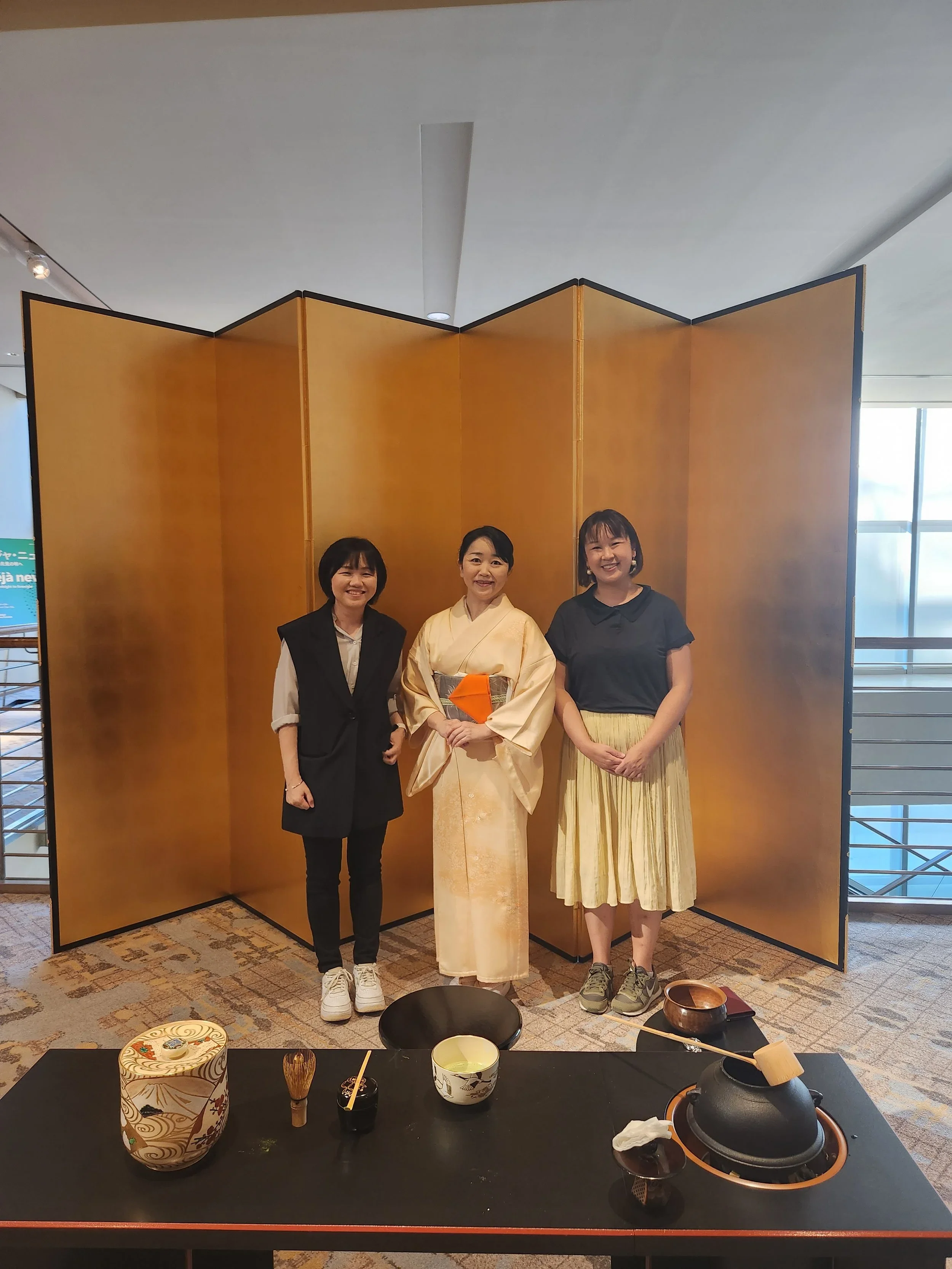 Three woman stand in front of a golden folding screen. One woman dressed in a kimono.