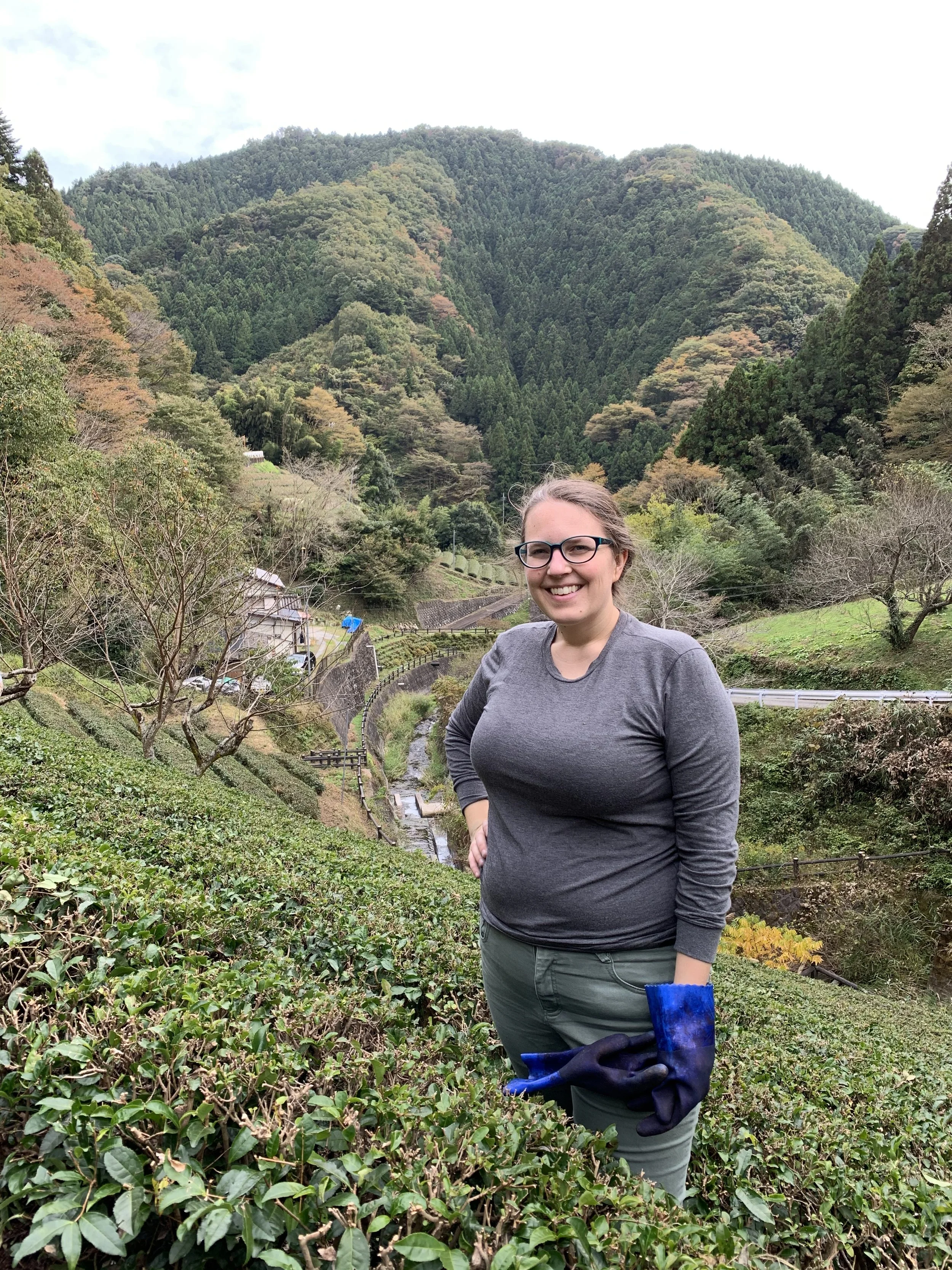 A woman with glasses standing in a lush green hilly landscape with trees and a small creek, smiling at the camera during daytime.