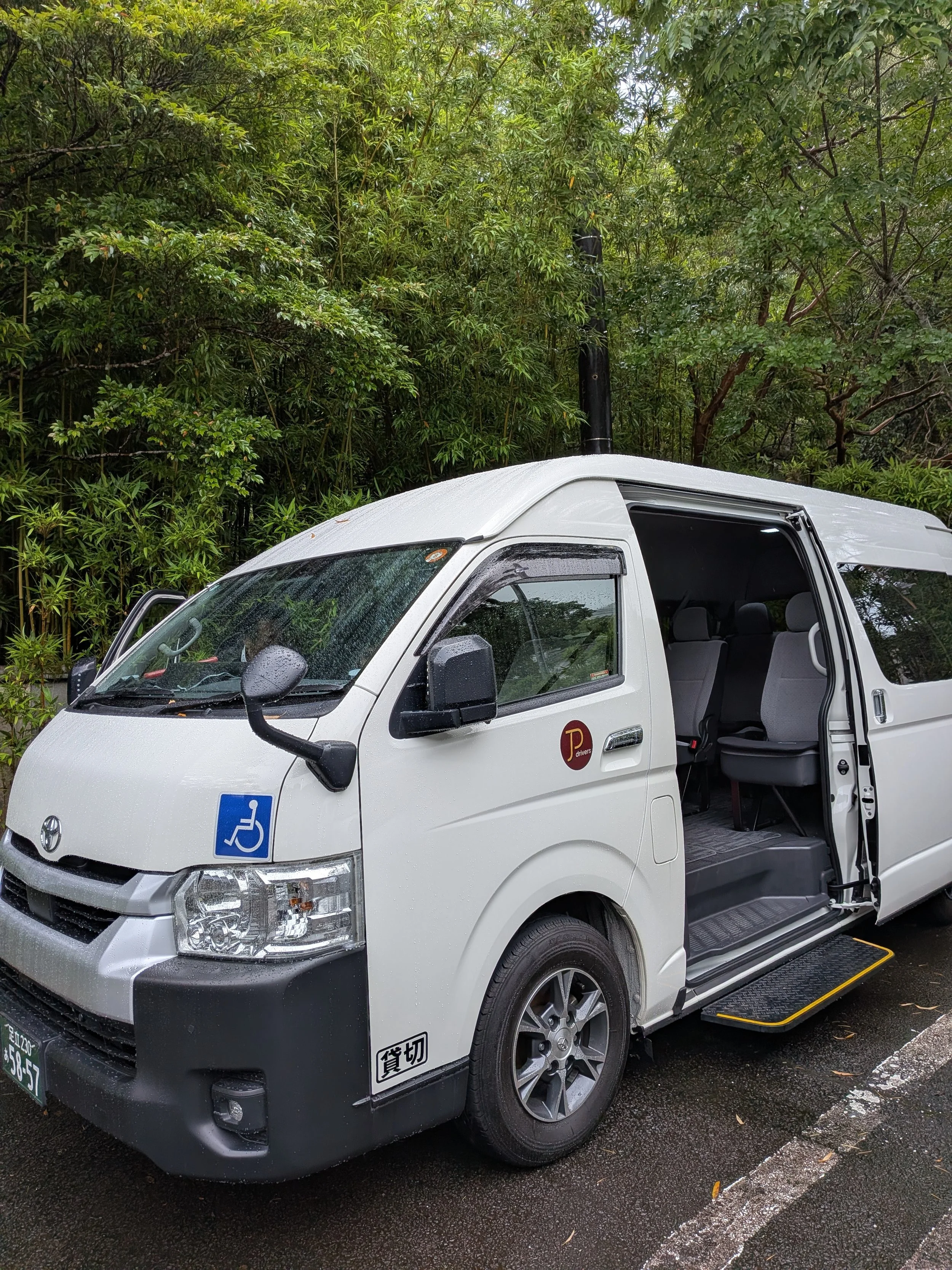 A white TokudAw Japan Private Drivers van parked in front of trees with an accessible sticker on the front side.