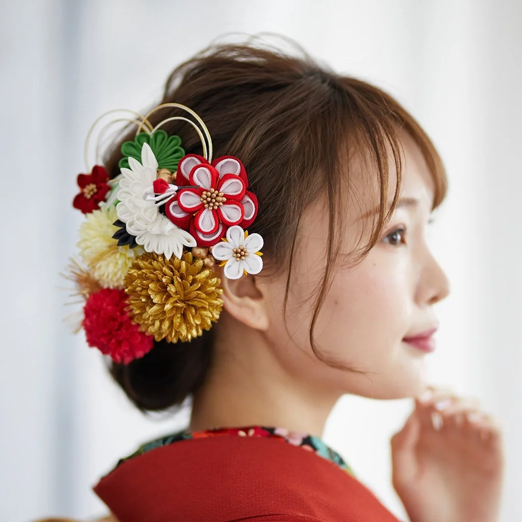 A side profile headshot of woman wearing a red kimono and an updo with a large white, gold, and red floral decoration in her hair