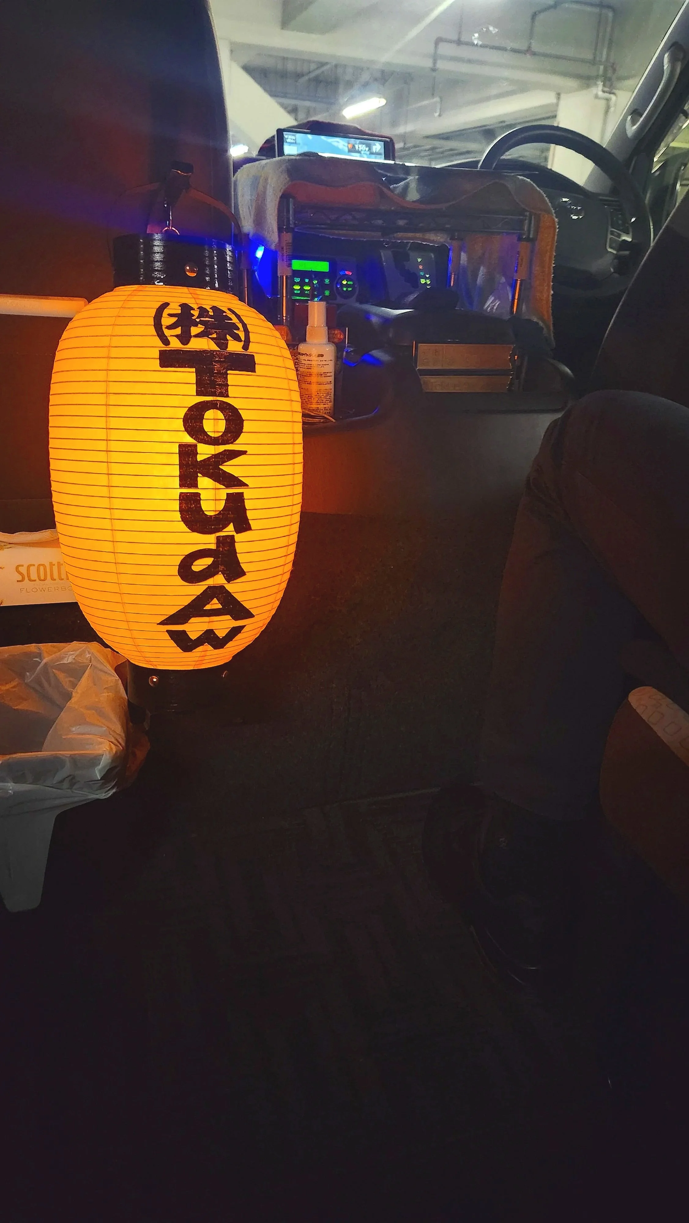 Interior of a vehicle with a Japanese paper lantern displaying Japanese characters, various electronic devices, hand sanitizer, and personal items on the dashboard, in a dimly lit parking garage.