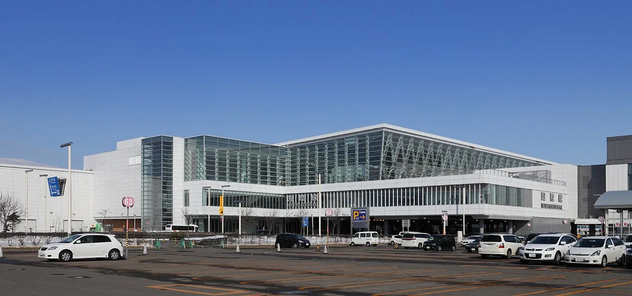 Image from Wikipedia, used under the Creative Commons Attribution-Share Alike 3.0 Unported license. Image shows a white and glass geometric building taken at an angle on the corner of the building with a parking lot in front with several white cars.