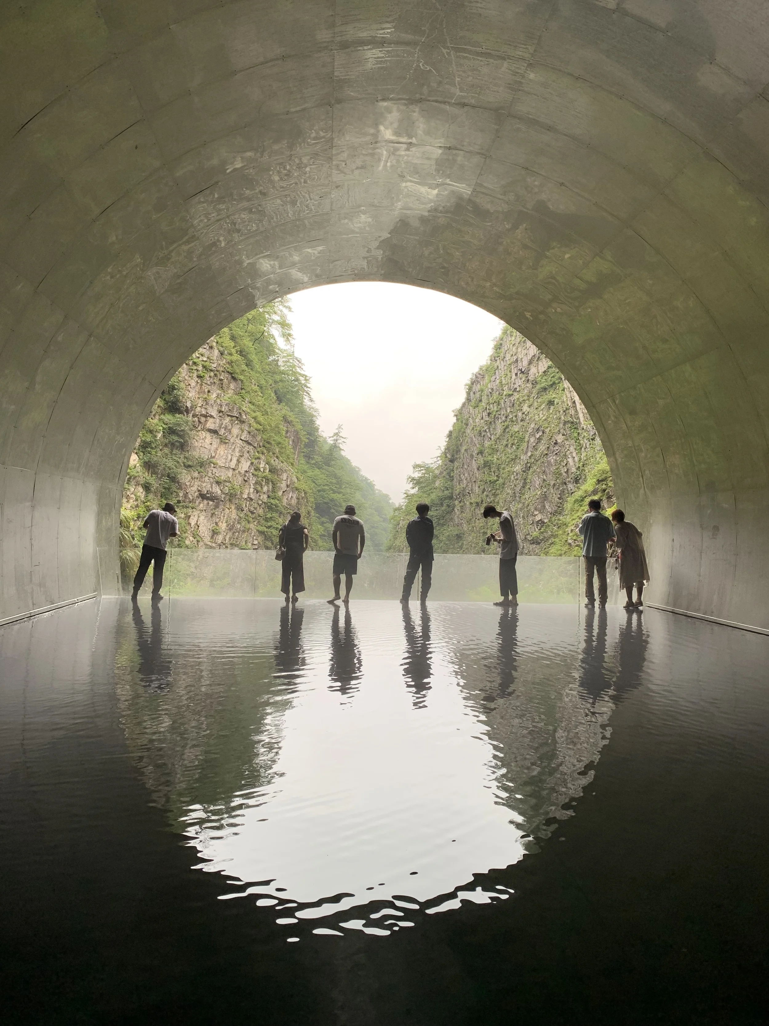 View of the end of a tunnel framing a nature scene. Inside the tunnel is reflective water, figures standing at the end of the tunnel are reflected on the water. A famous art locaiton in Niigata  prefecture, Japan.