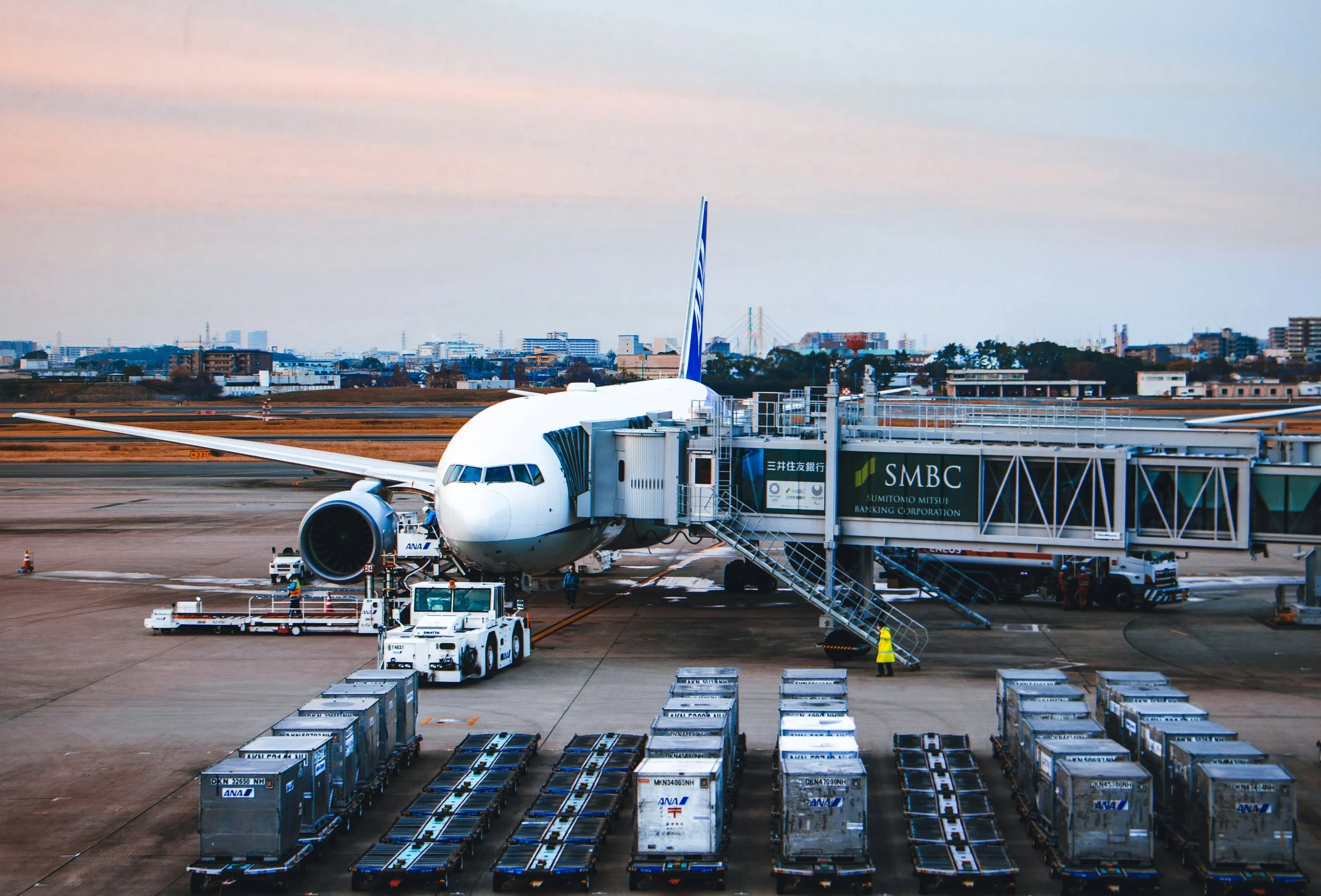 An airplane parked at an airport gate with a jet bridge attached. The scene shows airport tarmac areas with baggage carts and ground crew vehicles nearby, and a city skyline with buildings in the background under a partly cloudy sky.