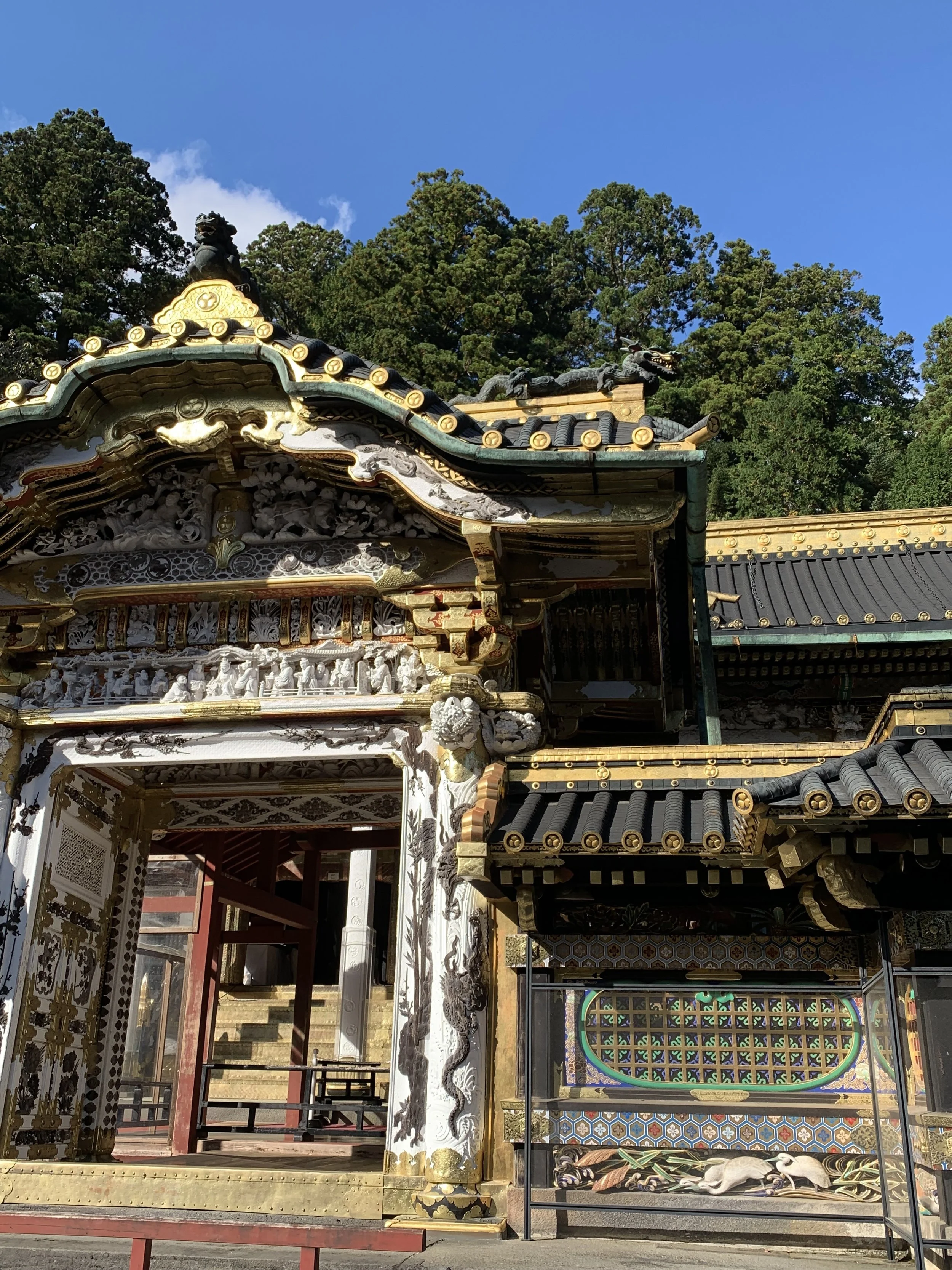 Close-up of a traditional Japanese building with ornate wood carvings, dragon sculptures on the roof, and decorative tiles, surrounded by green trees and a clear blue sky.