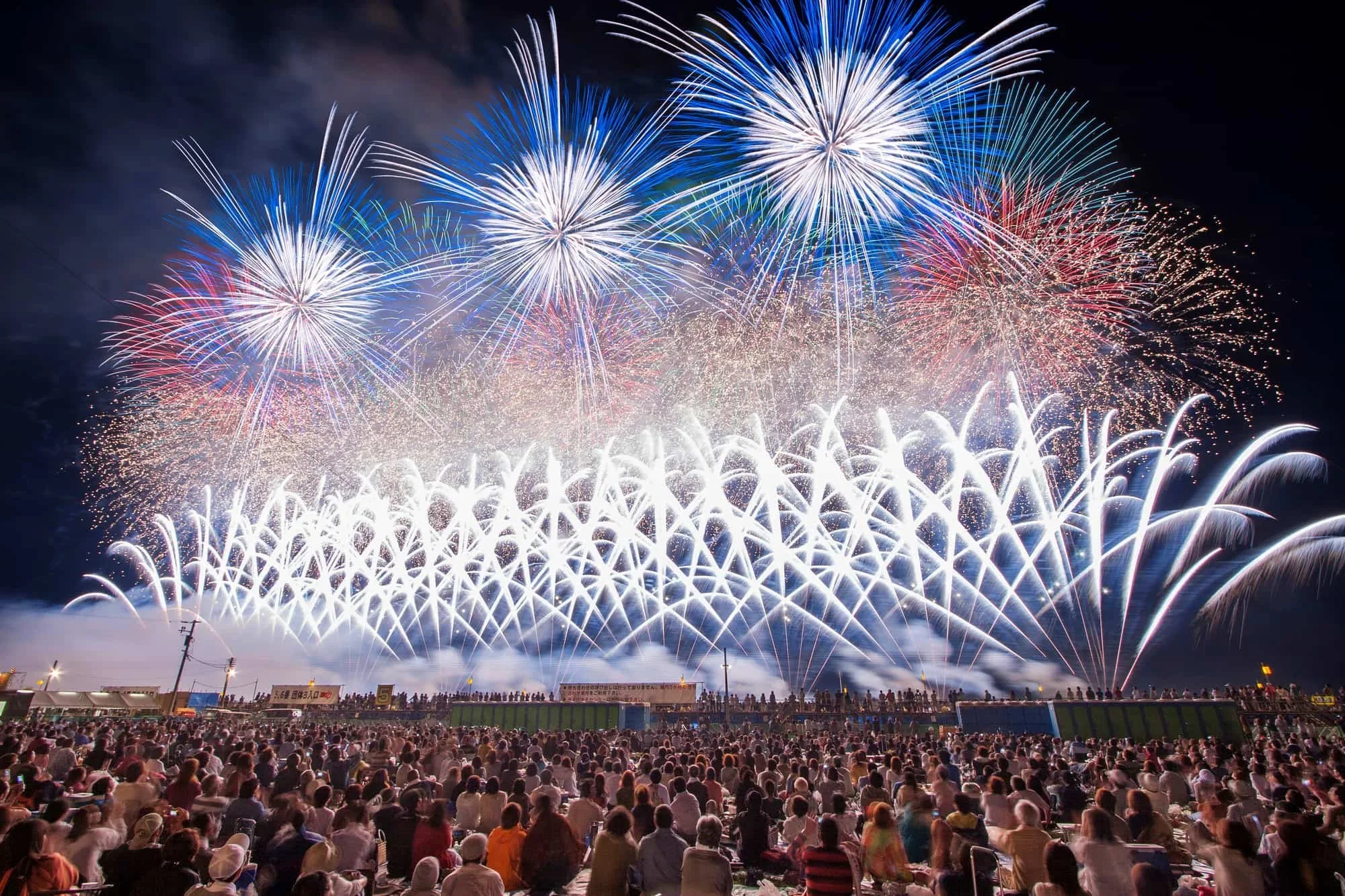 Crowds watch the Oomagari Fireworks Festival at Akita Prefecture with 3 big bursts of blue and white and many sparkling fireworks below it.