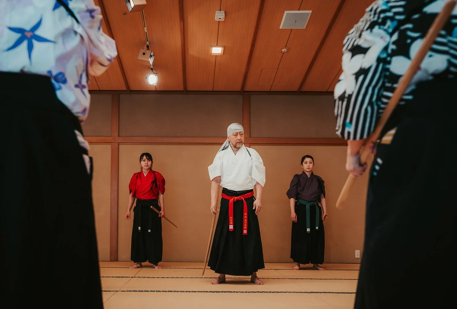 Samurai trainer stands in the middle of a tatami mat room with a weapon in hand and two students behind him.