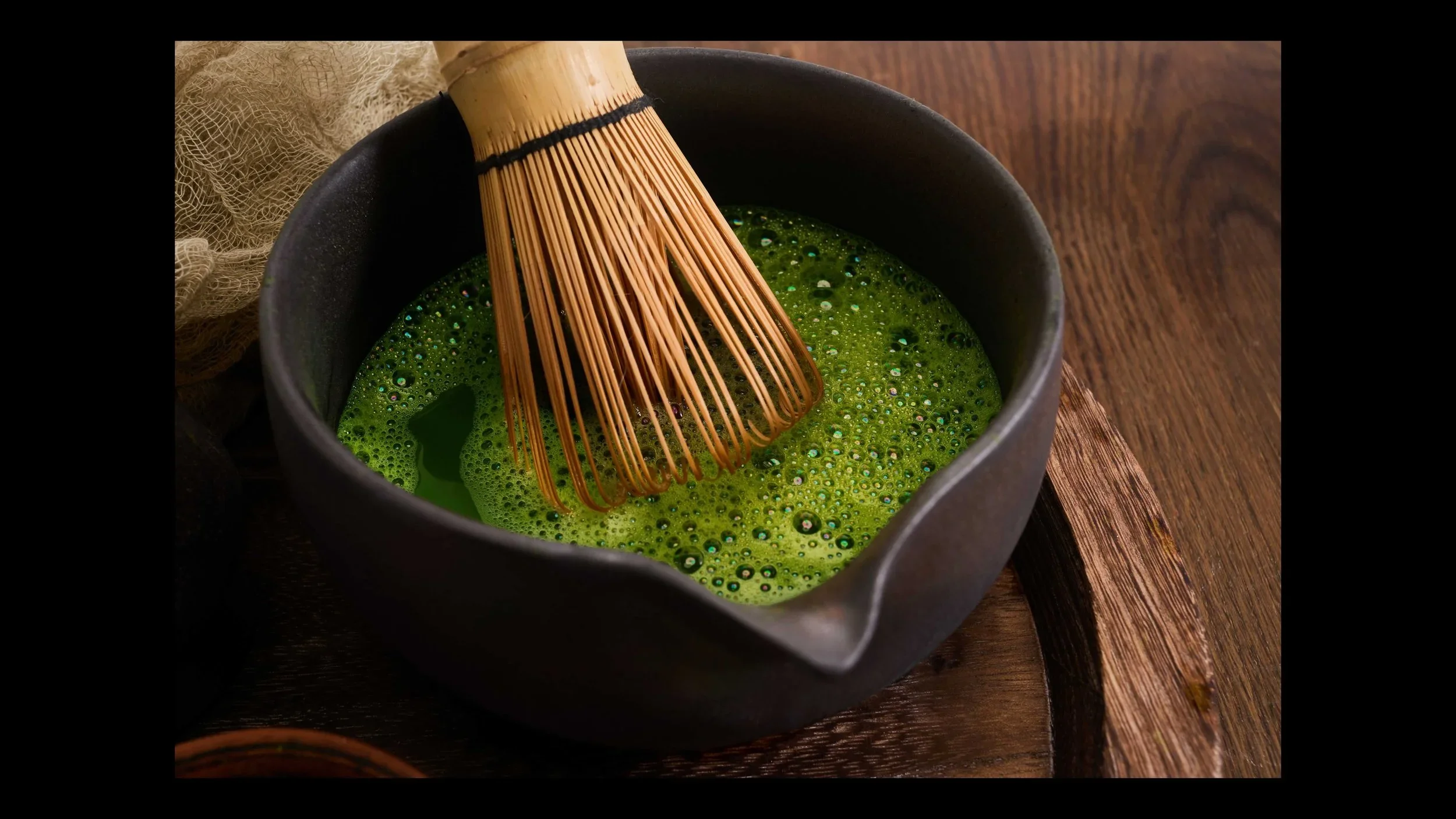 An image of frothed matcha with the bamboo whisk resting inside a black cup on top of a wooden tray.