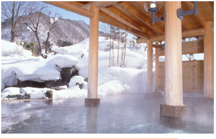 A covered wooden warm onsen amongst a winter scene. Onsen overlooks a snow covered rock garden and steam rises from the onsen water.