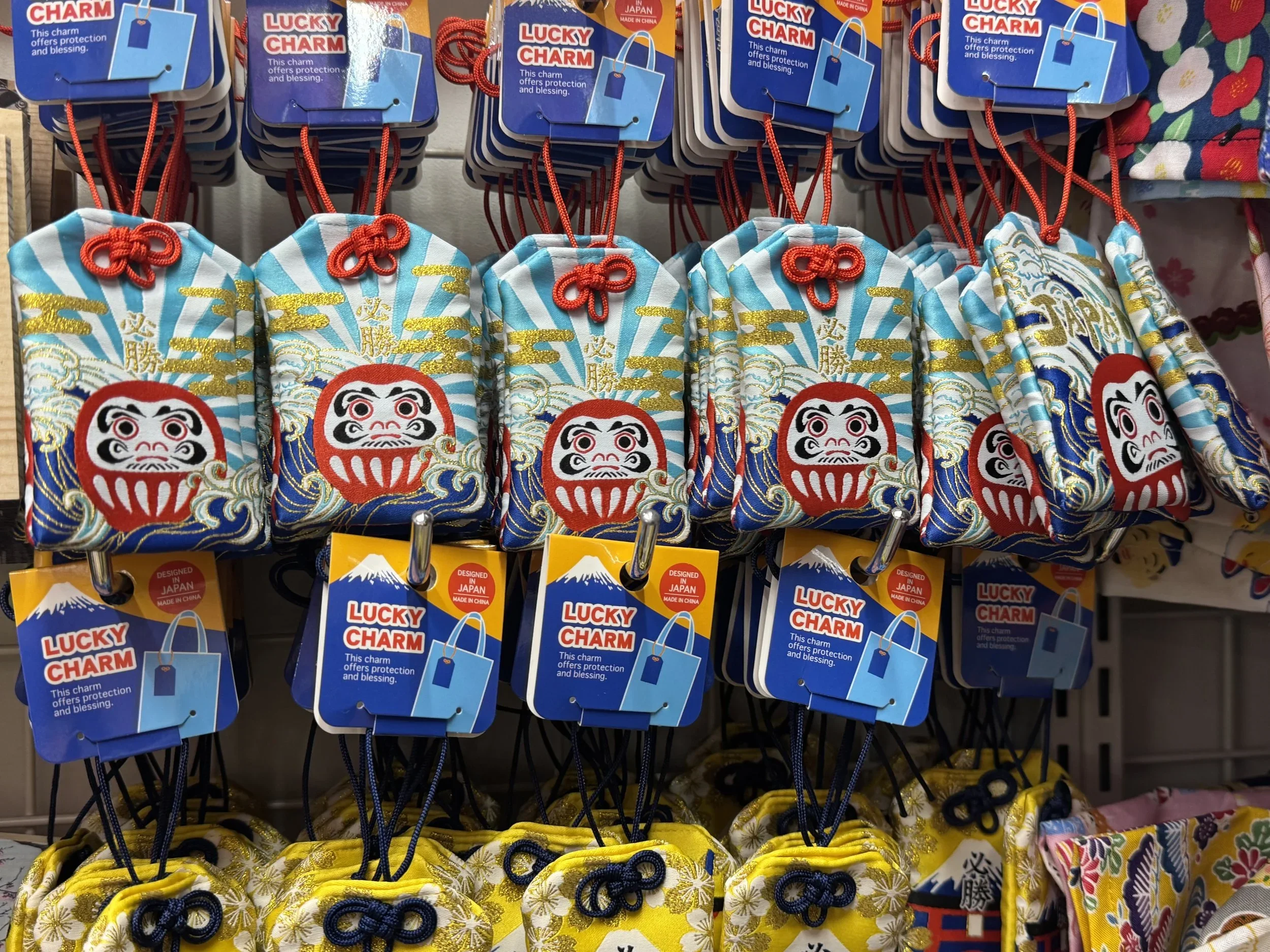 Japanese fabric pouch good luck charm called an omamori hangs from a hook featuring a red, round daruma doll, gold clouds, a wave, and blue and white sun-burst like streaks in the background.