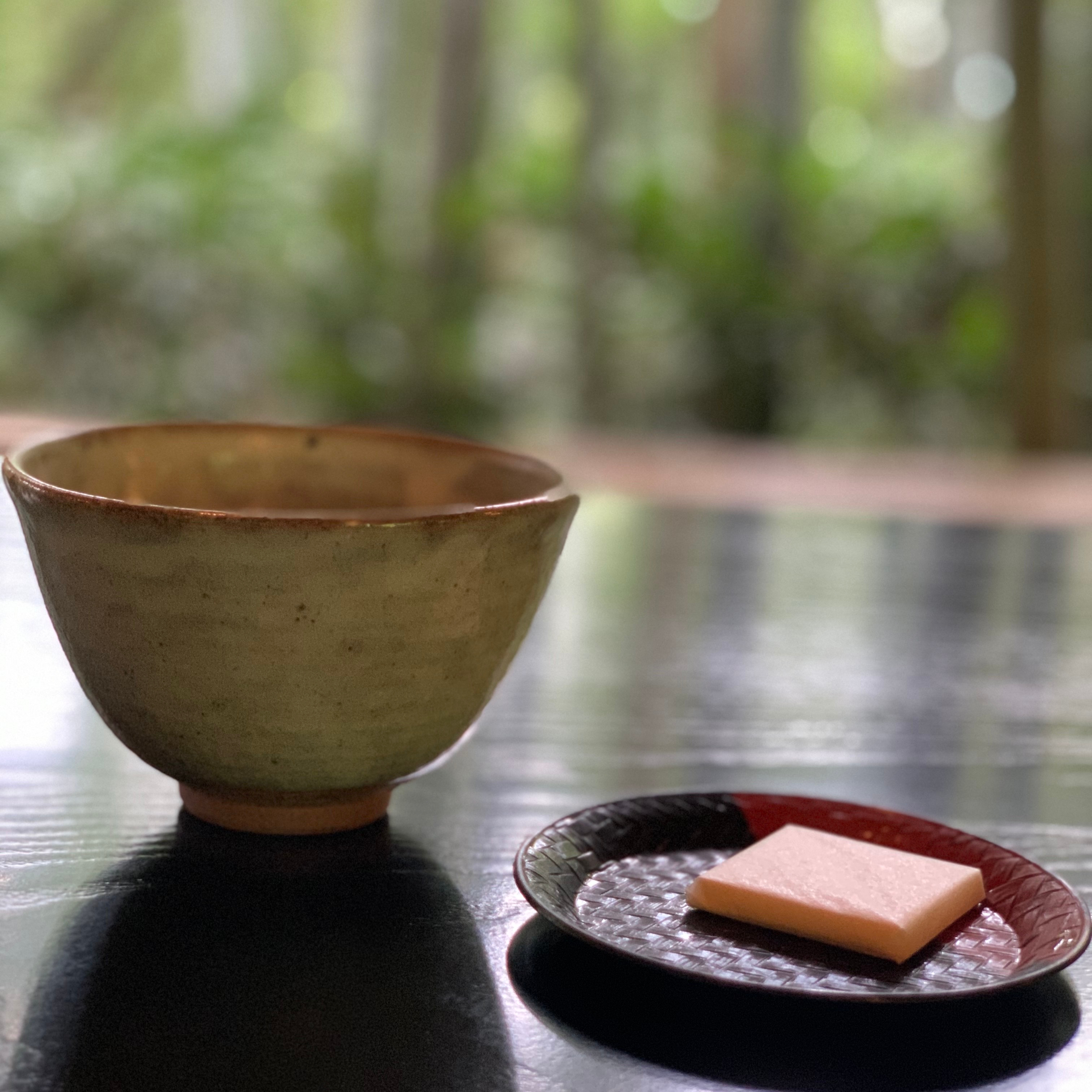 A small earthenware teacup sits next to a japanese sweet on a lacquerware plate on a table in a Japanese garden.