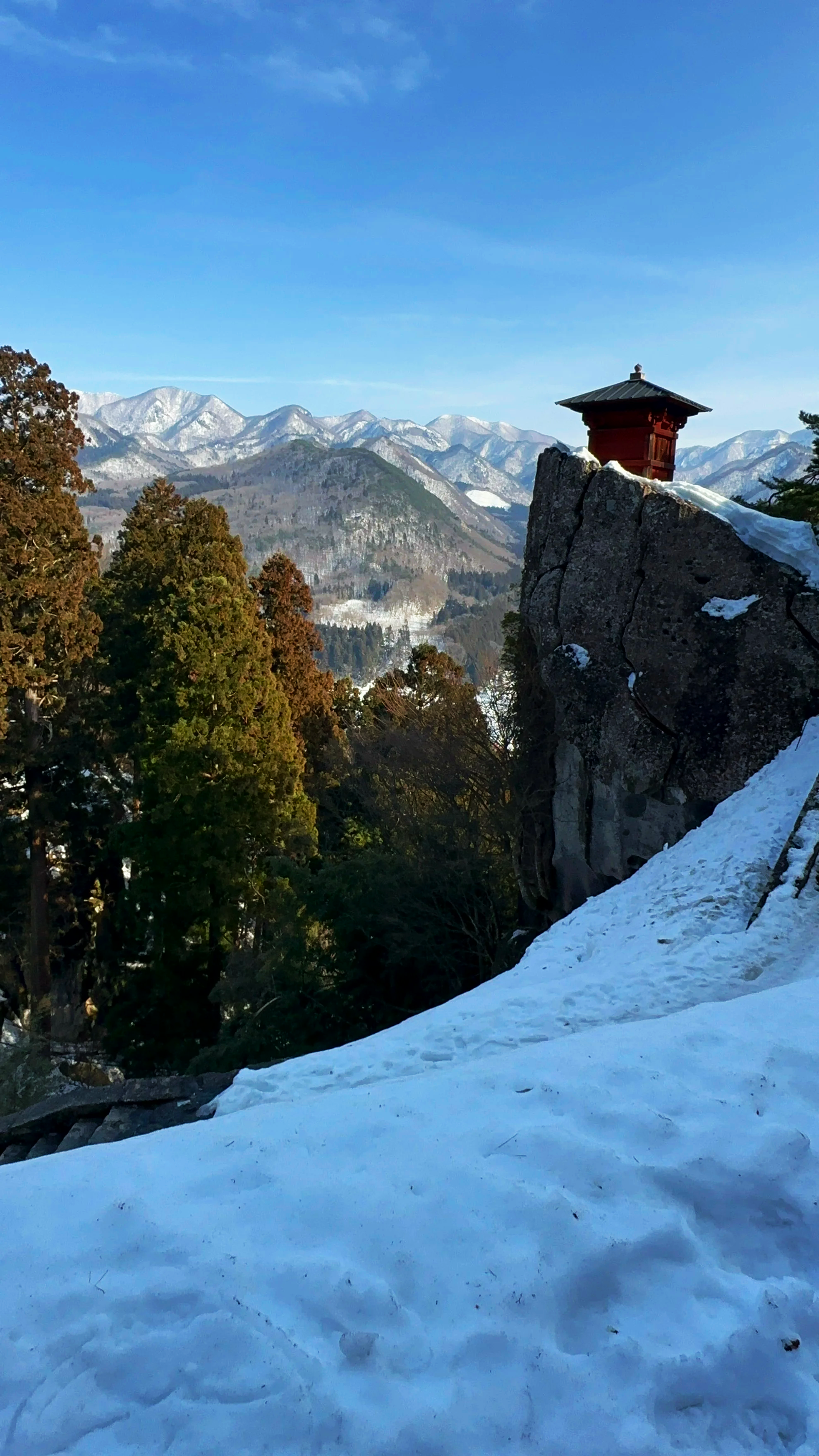 A snowy landscape with snow-covered ground in the foreground, green trees on the left, a rock with a small traditional building on top on the right, and mountain ranges with snow-capped peaks in the background under a clear blue sky.
