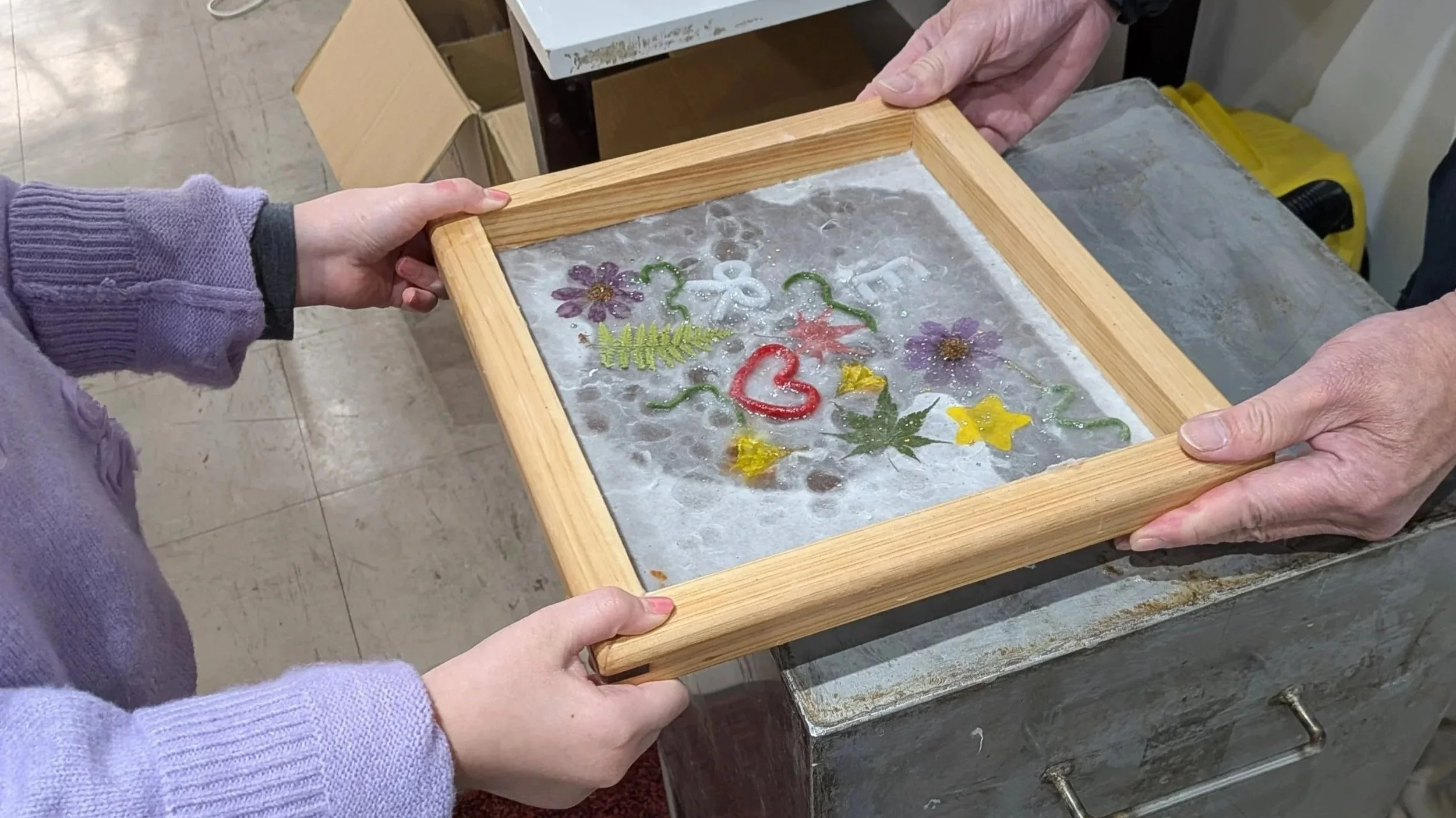 A child and older adult work together to sift paper pulp mixed with flower petals. w