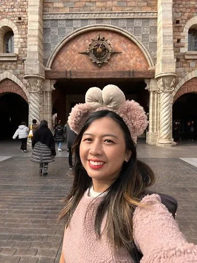 Portrait of a woman wearing pink and Shelly Mae ears with brown hair standing in front of a tiled archway at Tokyo Disney Sea.