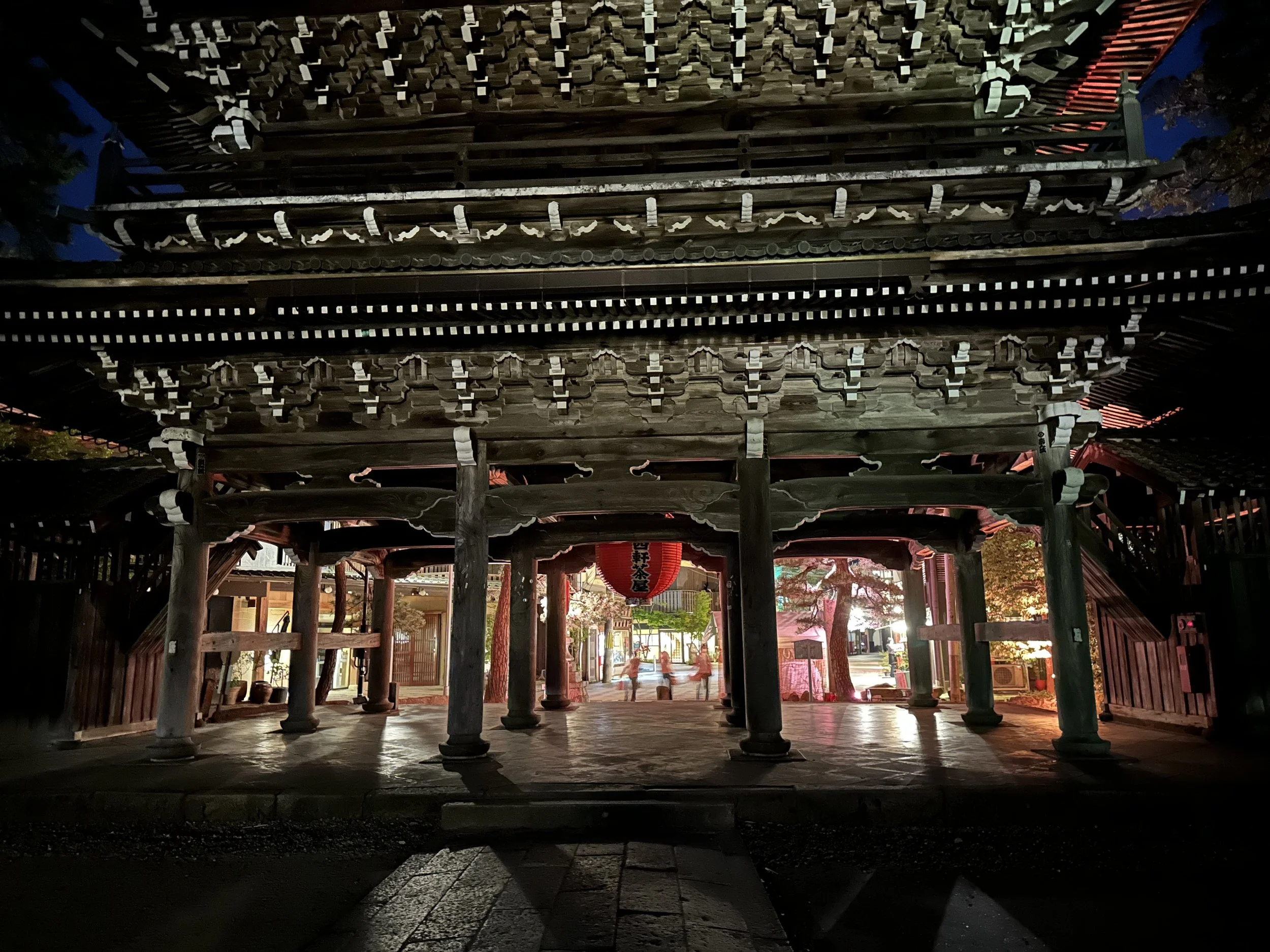 A traditional Japanese temple gate (torii) at night, constructed of dark wood with intricate architectural details, illuminated softly from below, with a red paper lantern hanging inside and a pathway leading inside.