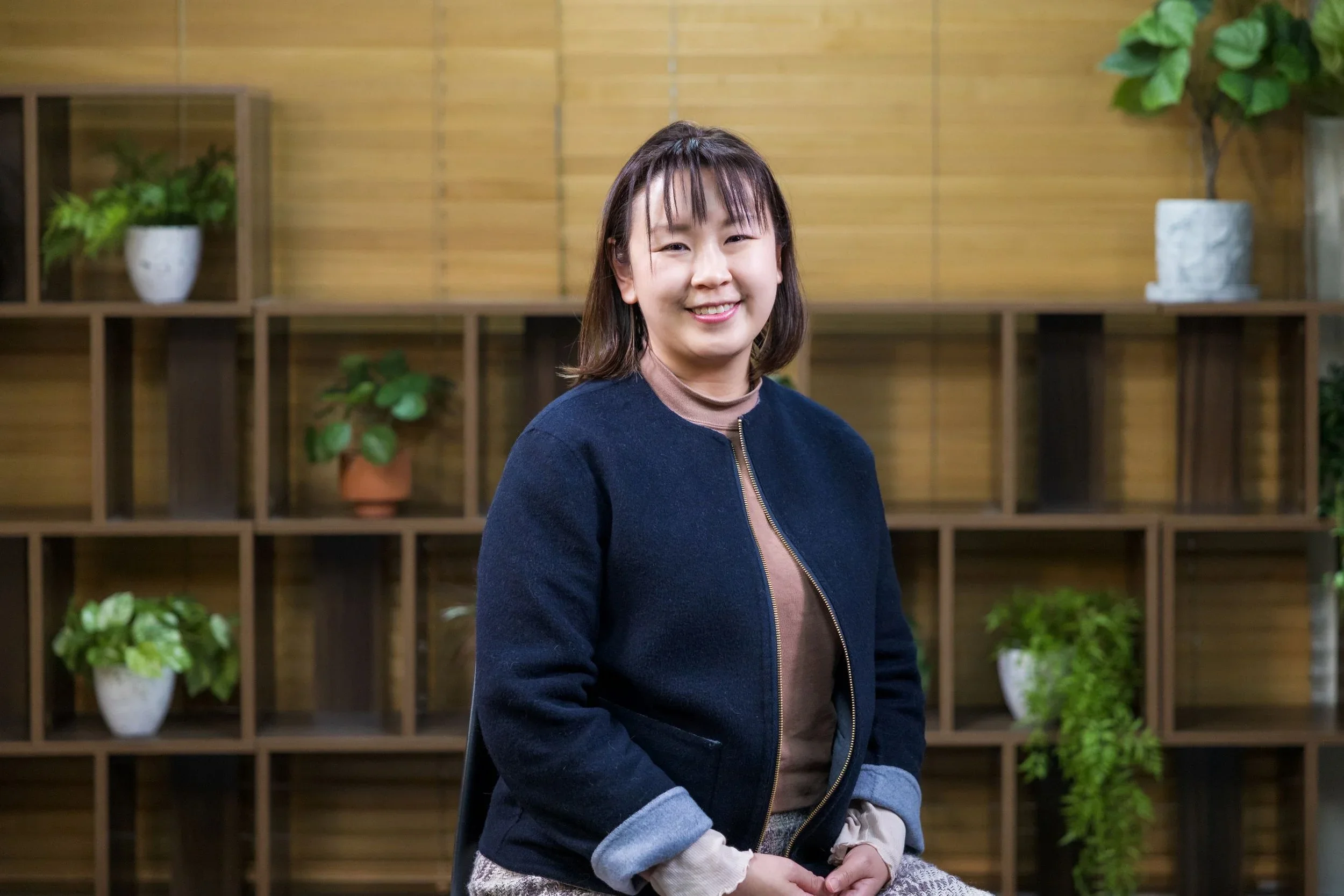A young woman with shoulder-length dark hair wearing a navy jacket and beige sweater sitting in front of a wooden shelf with potted plants.