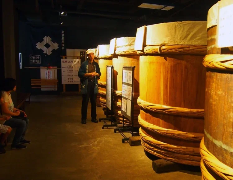 Man stands next to a row of 5 large wooden miso vats covered with white fabric on top and standing on a dirt floor.