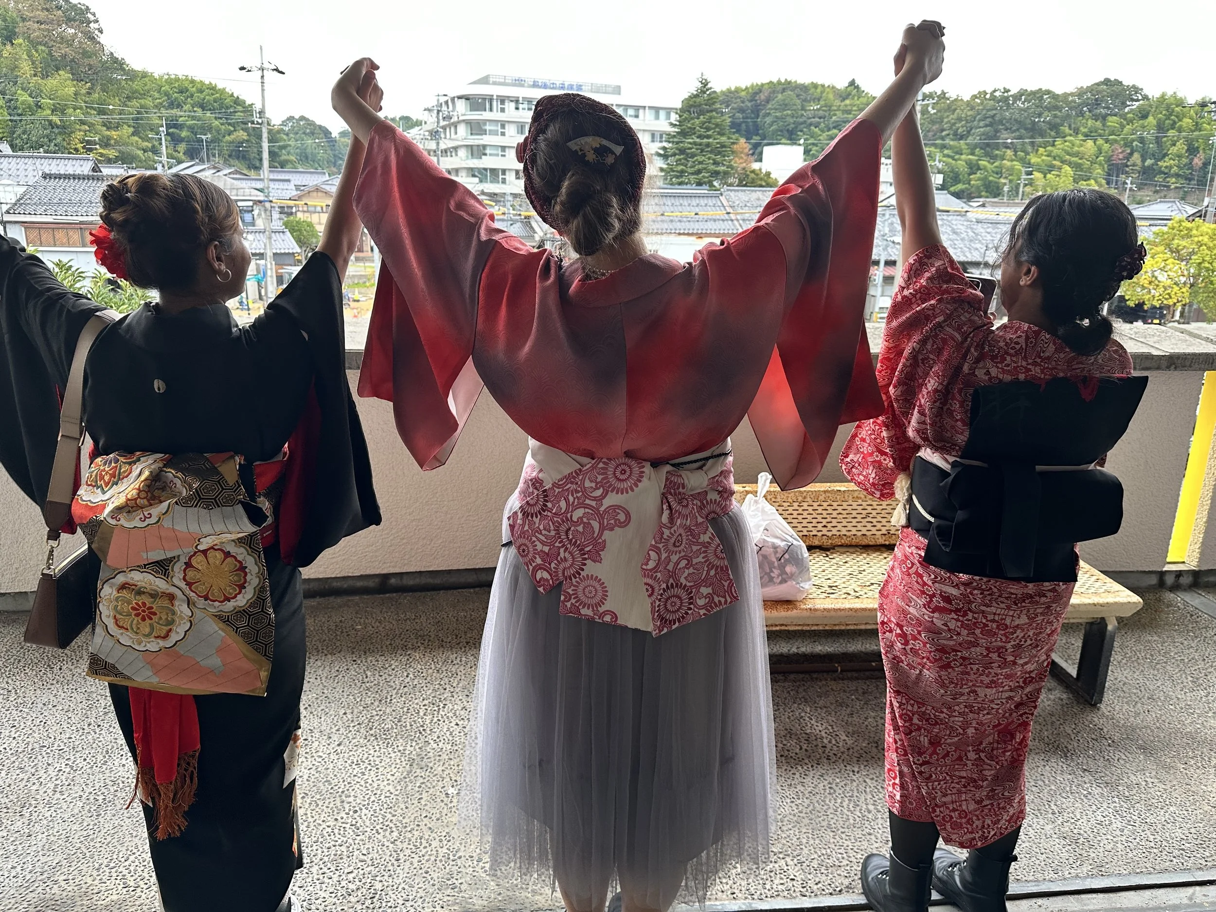 Three women in traditional Japanese kimonos holding hands in a circle outdoors, with a background of buildings and trees.