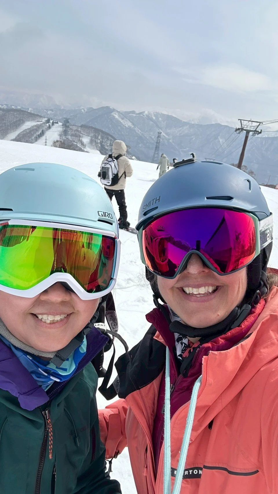 Two women in ski gear taking a selfie on a snowy mountain with ski lifts and mountain peaks in the background.