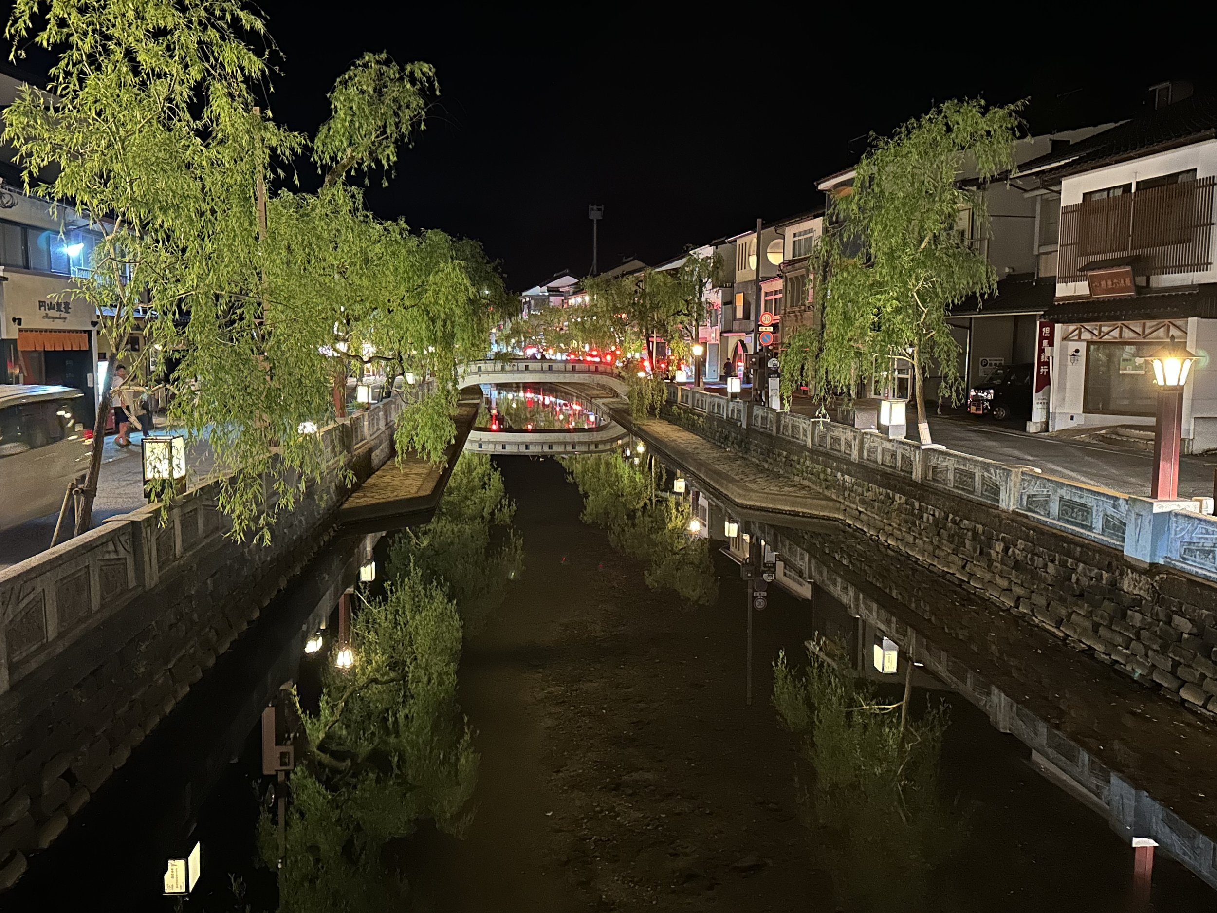 A canal lined with evenly spaced trees on either side and a stone wall and bridge with two story Japanese homes on either side at night.