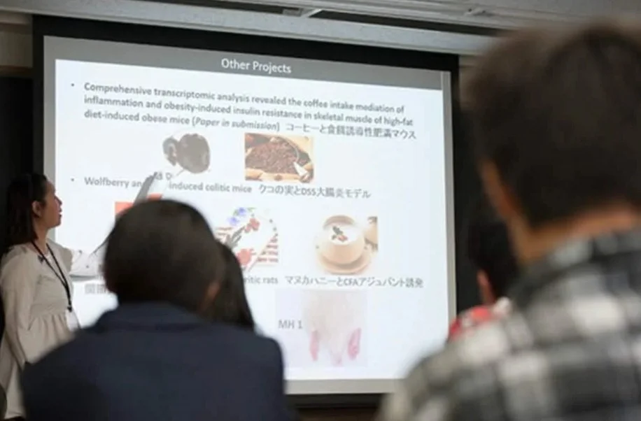 A woman is giving a presentation in a classroom or conference room, with several seated attendees. The presentation slide in Japanese discusses research projects involving mice models and includes images of mice and food.