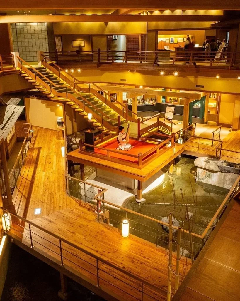 The lobby at Ookawaso that resembles a scene from Demon Slayer and features intertwining stairs with warm wood and an interior Japanese garden with rocks and water..