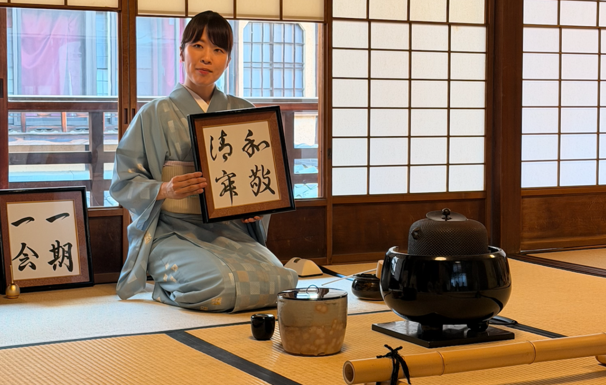 A woman in a blue kimono holds a framed calligraphy art and sits behind a tea ceremony tea pot and utentsils.