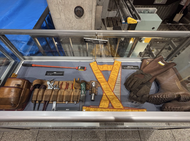 Tools used for building the Tokyo Metro Tunnels are on display in a small glass case. Tools include a tool belt, a reflective vest, boots and gloves.