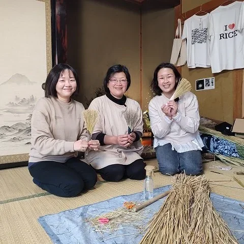 Three women sit in an traditional Japanese home with mini brooms and an I love rice  tshirt hanging in the background.
